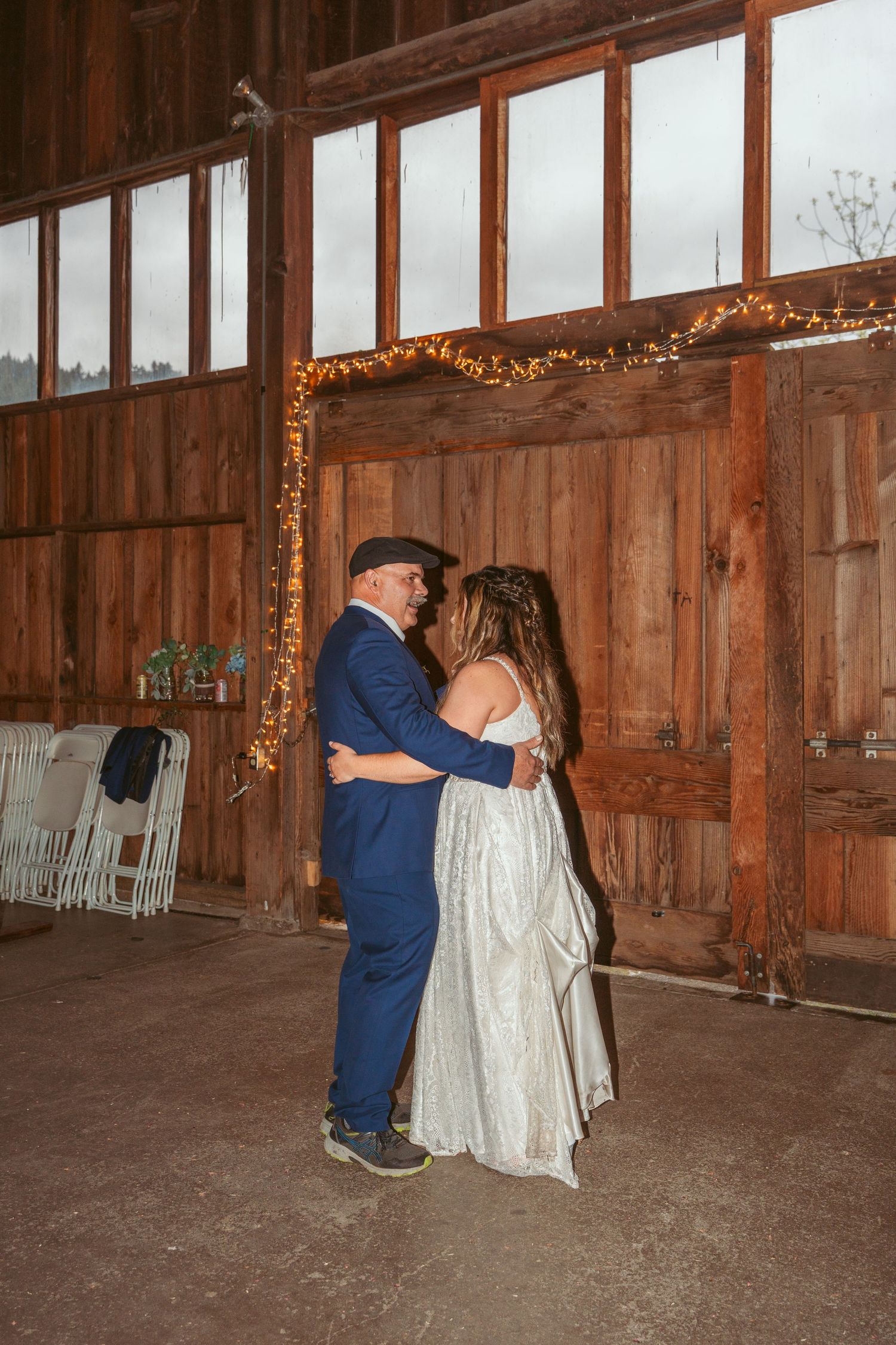 A couple embraces during their dance in a rustic barn with windows and string lights.