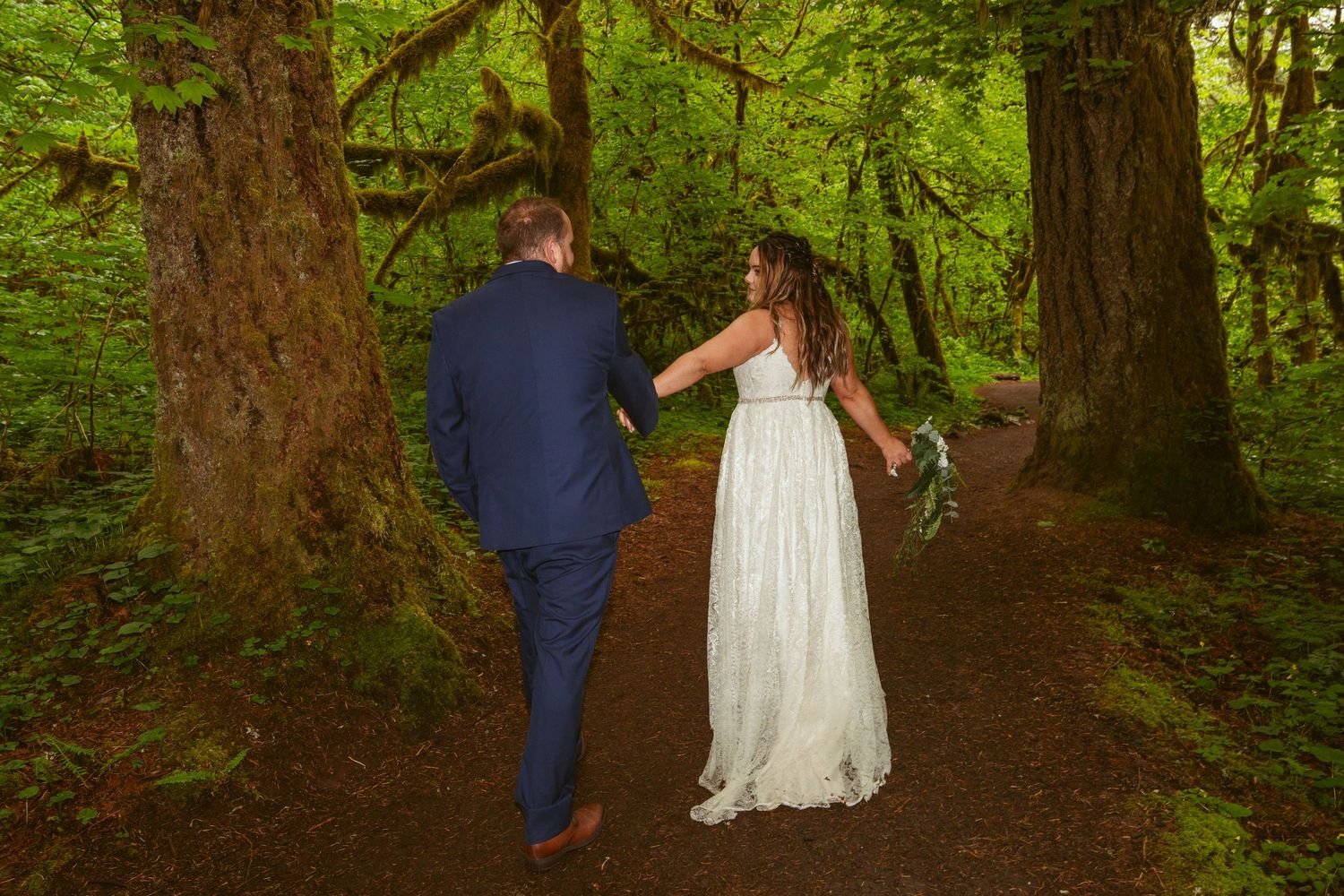 Newlyweds stroll together along a winding dirt path through a verdant forest.