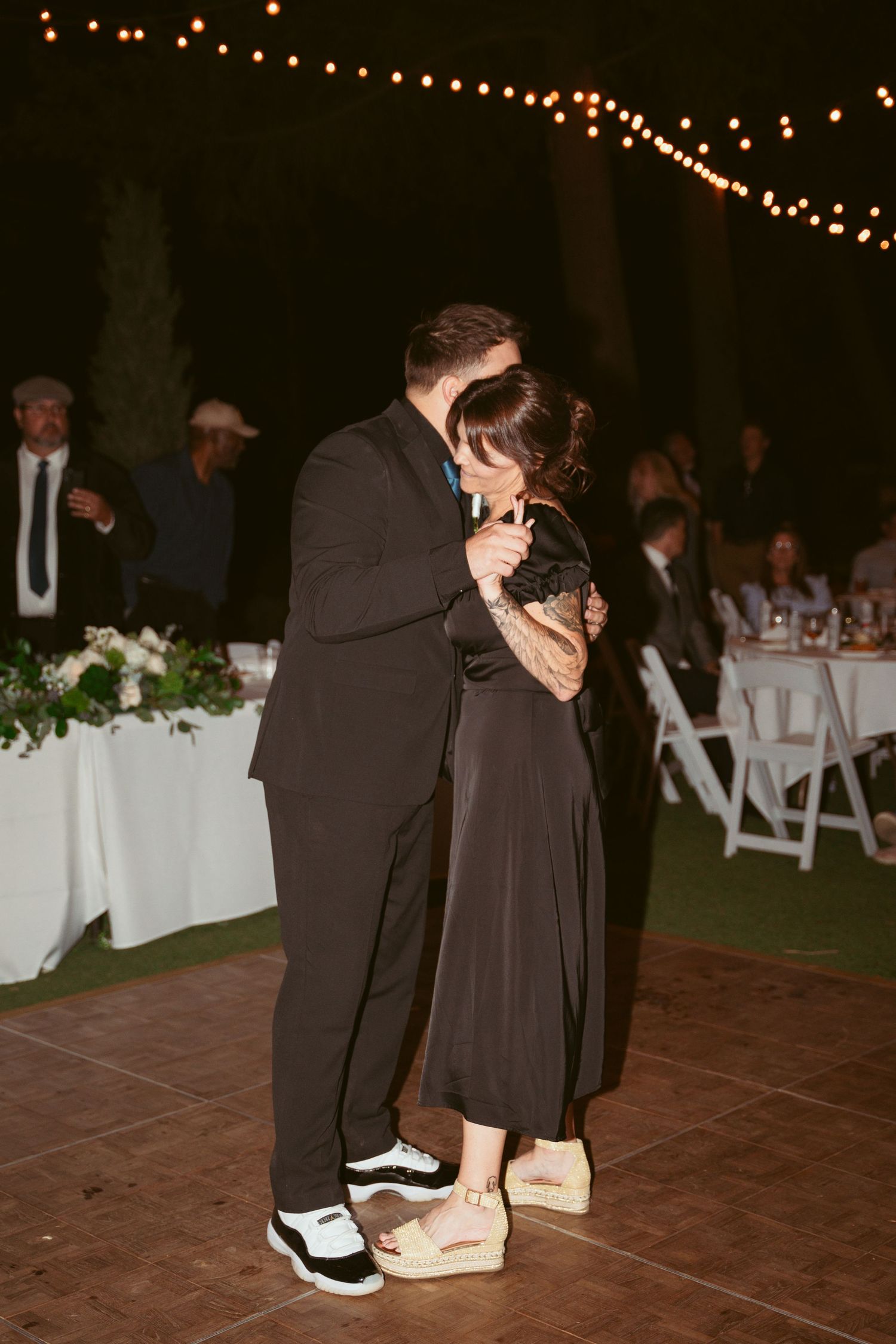 Couples slow dancing on a wooden floor with white reception tables in the background.