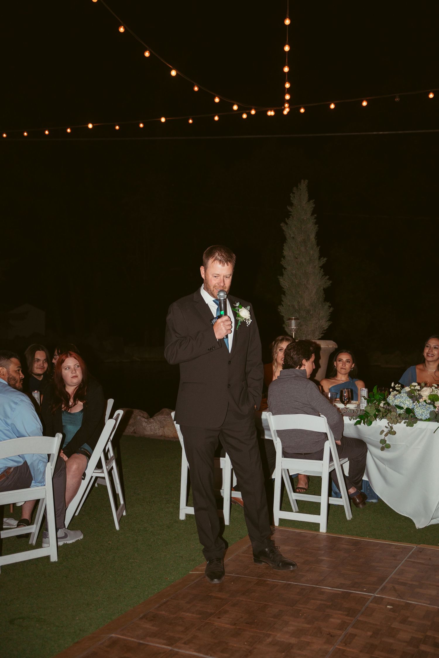 Wedding guests seated at tables with white chairs during an outdoor evening reception.
