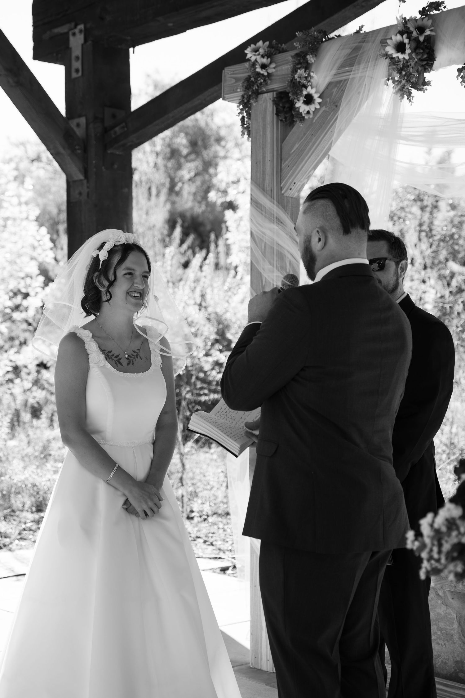 Wedding couple exchanges vows beneath a wooden pergola with autumn foliage in the background.