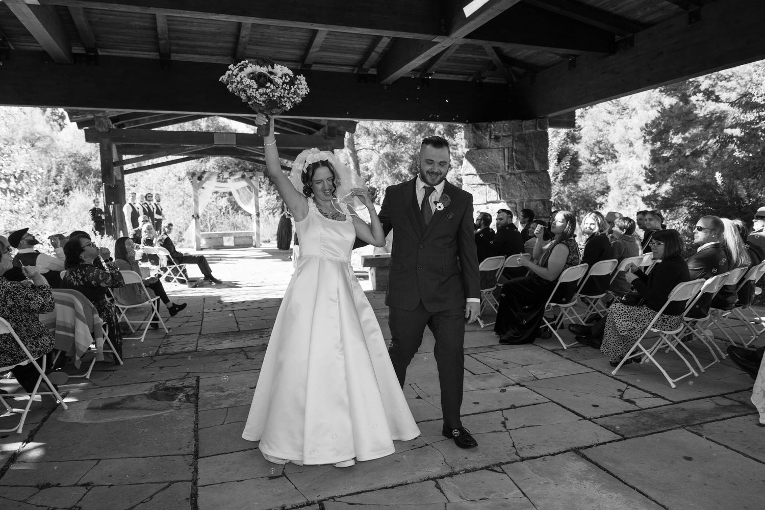 Black and white panorama of a wedding celebration under an open-air pavilion structure.