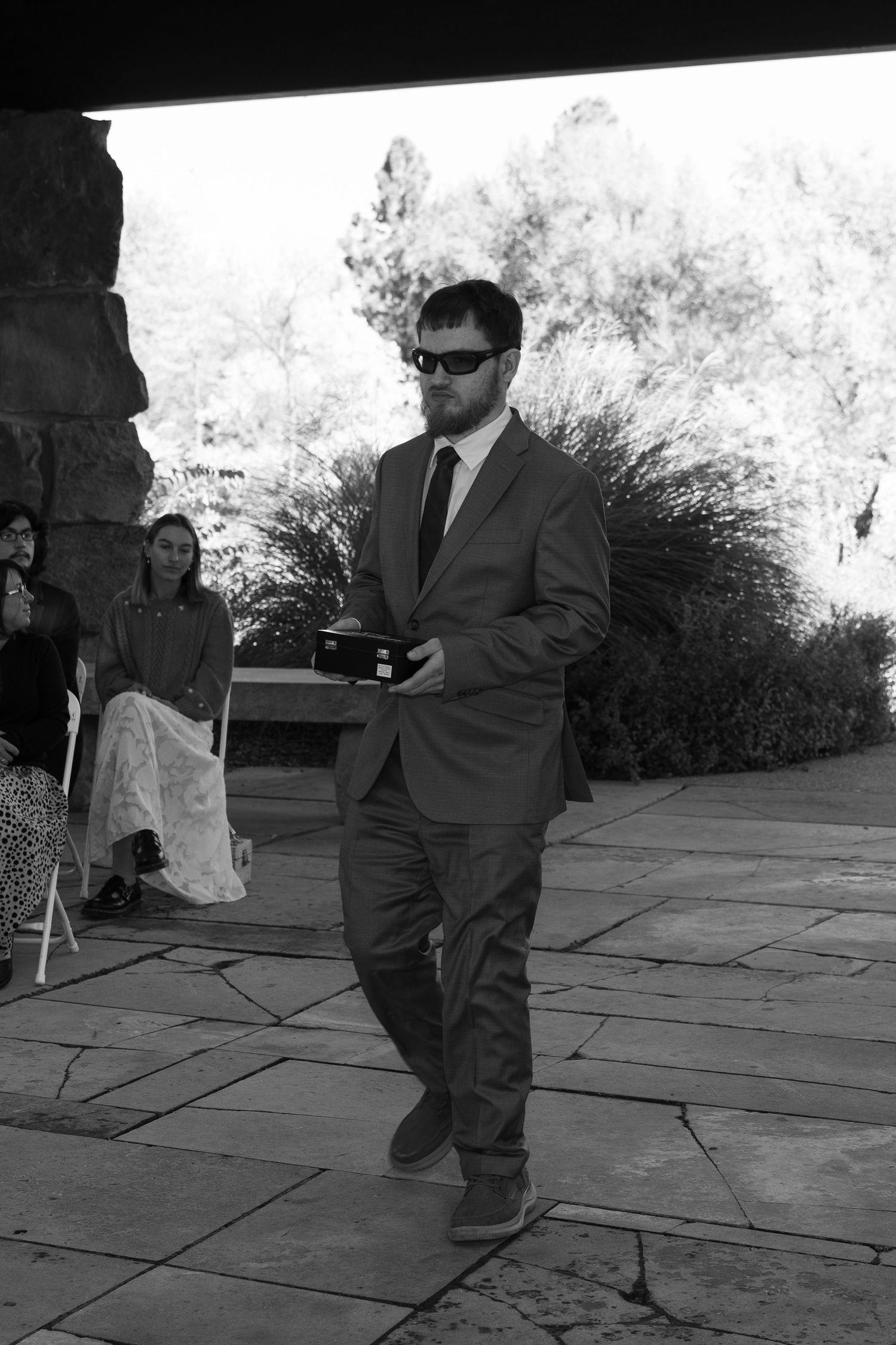 Black and white photo of attendant in suit and sunglasses walking during outdoor ceremony.