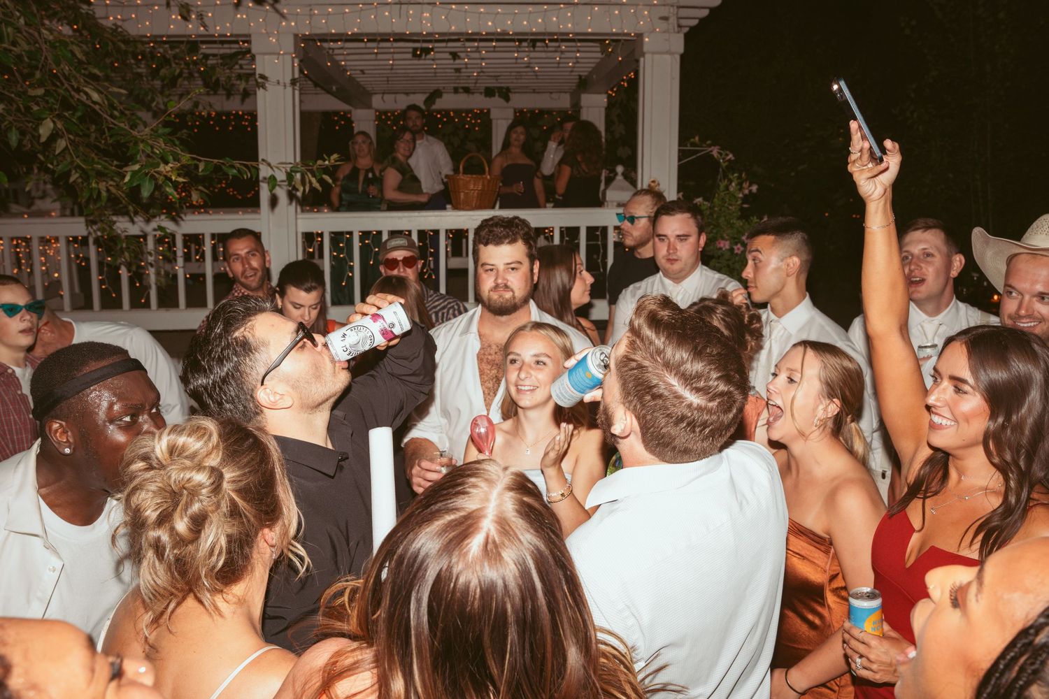 Crowded dance floor at an outdoor wedding reception with white pillars and string lights.