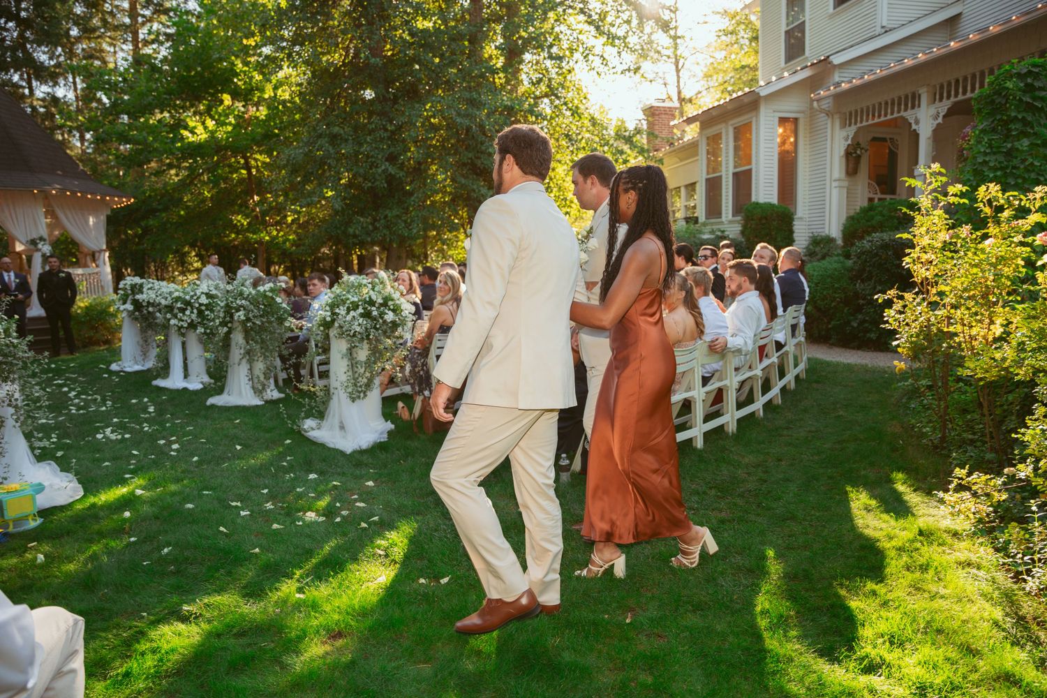 Summer wedding scene with attendees gathered on manicured lawn beside Victorian architecture.
