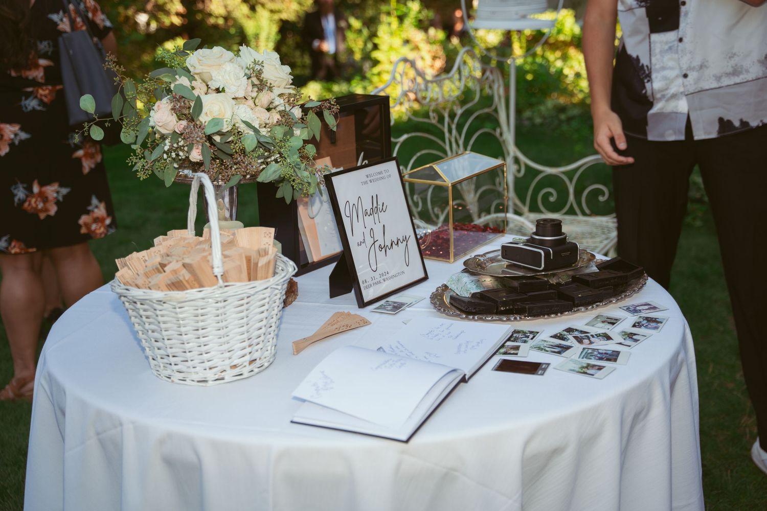 Elegant wedding dessert and gift table decorated with white linens and floral arrangements in an outdoor setting.