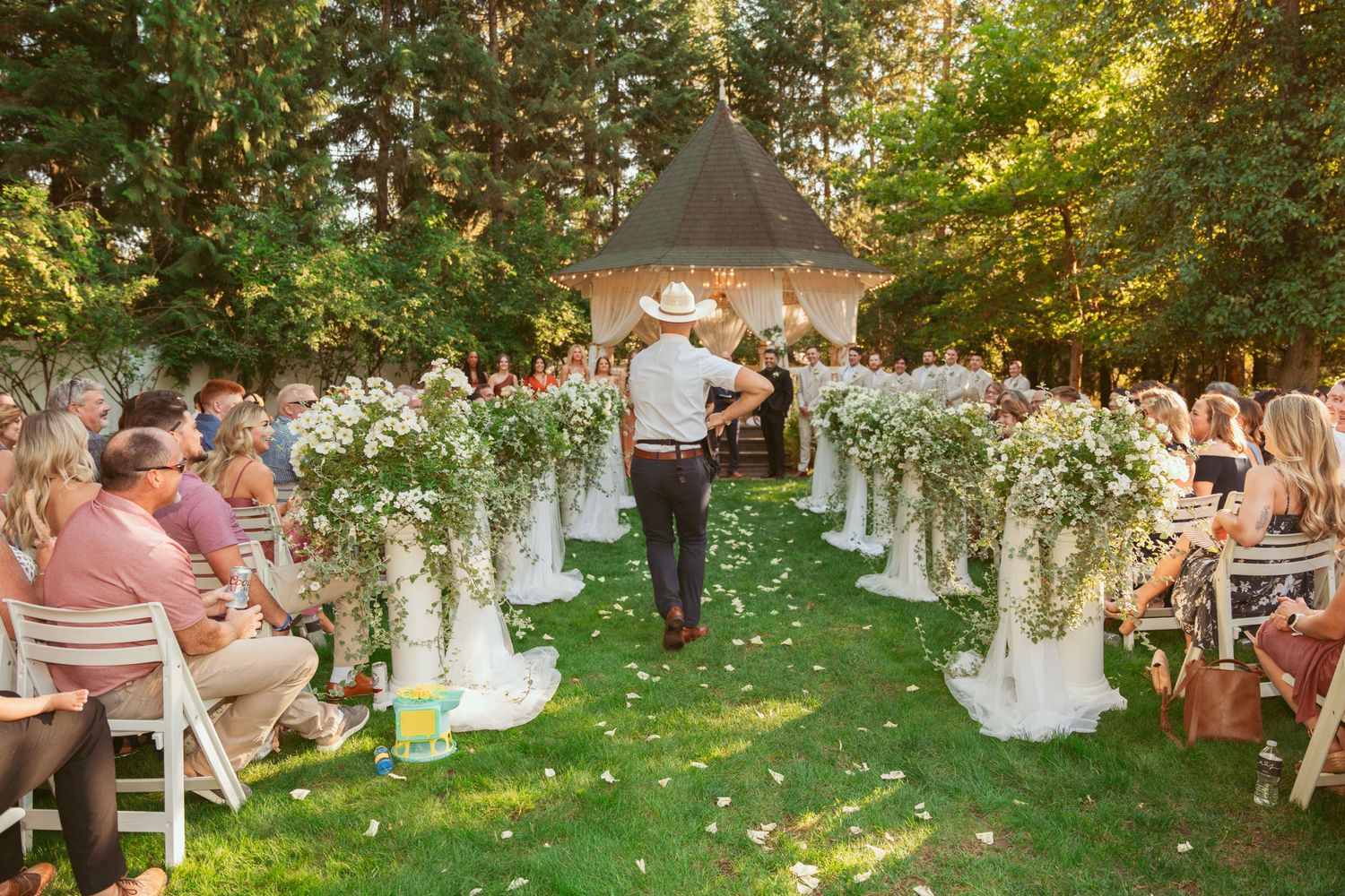 Outdoor garden wedding ceremony with white chairs and floral arrangements under trees at sunset.