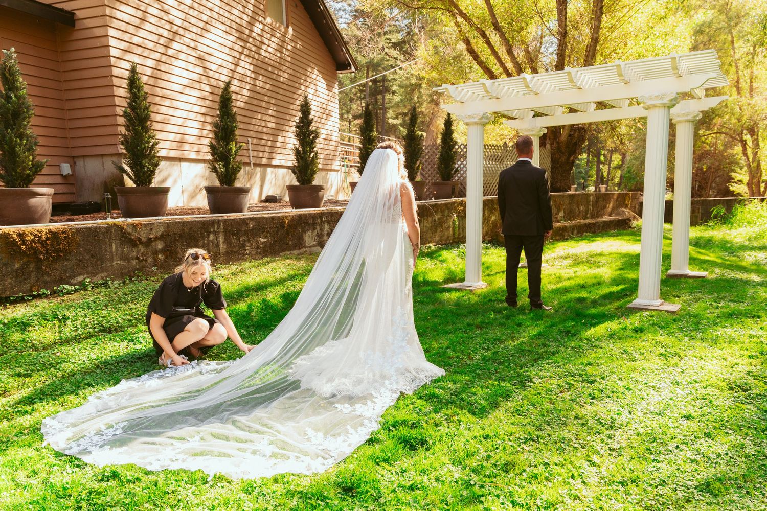 A dramatic cathedral veil trails behind a wedding dress as someone adjusts it under a garden pergola.