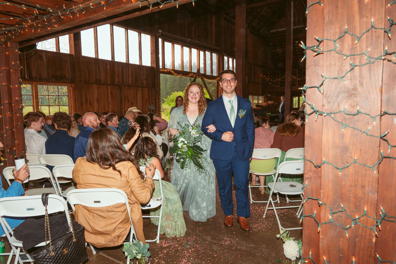 Wedding processional walking down aisle in barn venue with wooden walls and string light decor.