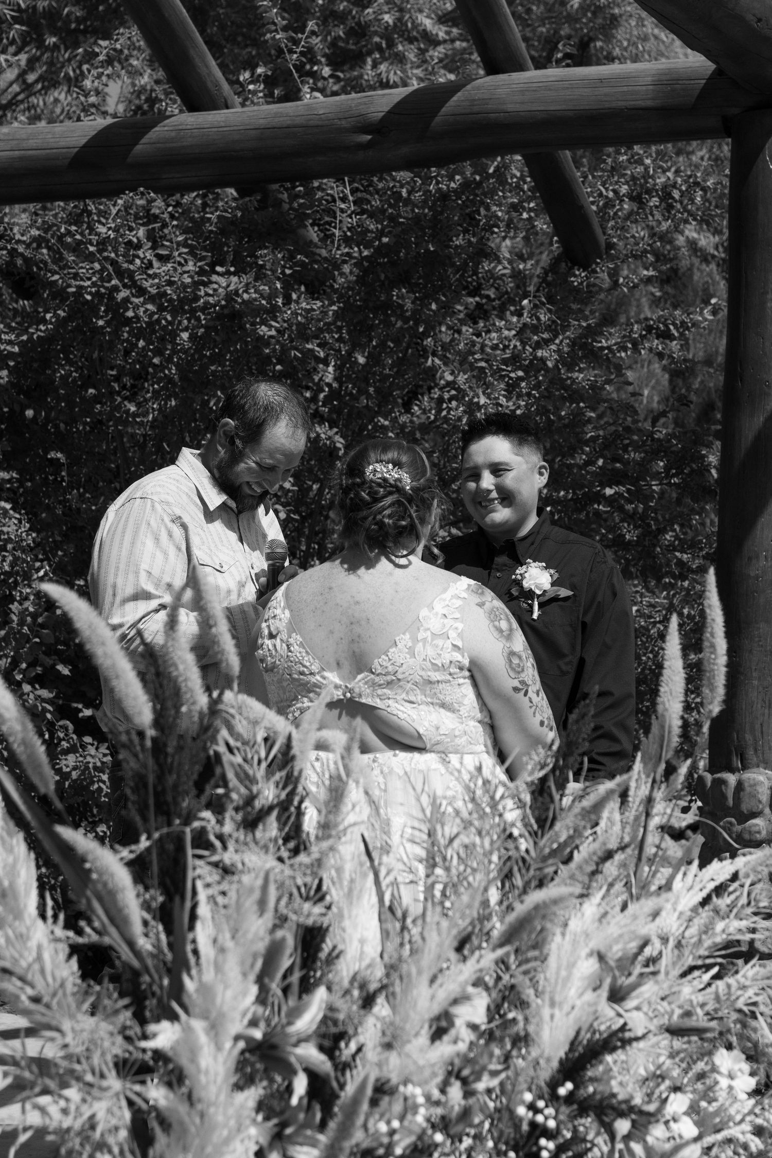 A group gathers for an intimate moment during a wedding ceremony under a wooden pergola with decorative flowers.