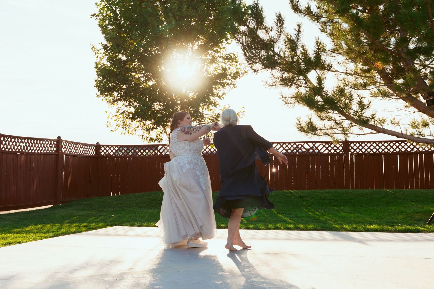 Newlyweds dance together on a white outdoor platform with sunlight streaming through pine trees.