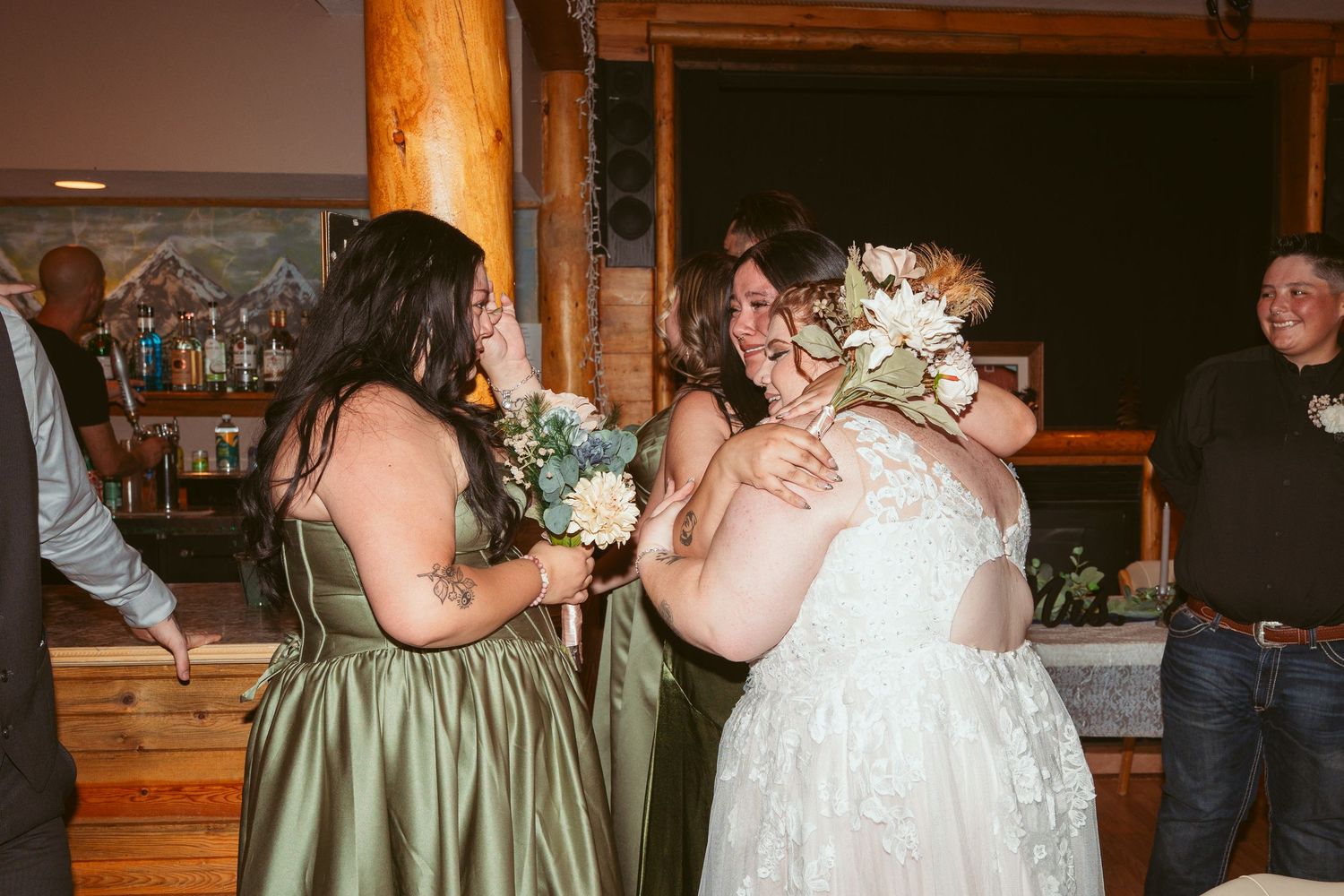 Emotional wedding celebration moment with people embracing while holding bouquets in a rustic wooden venue.