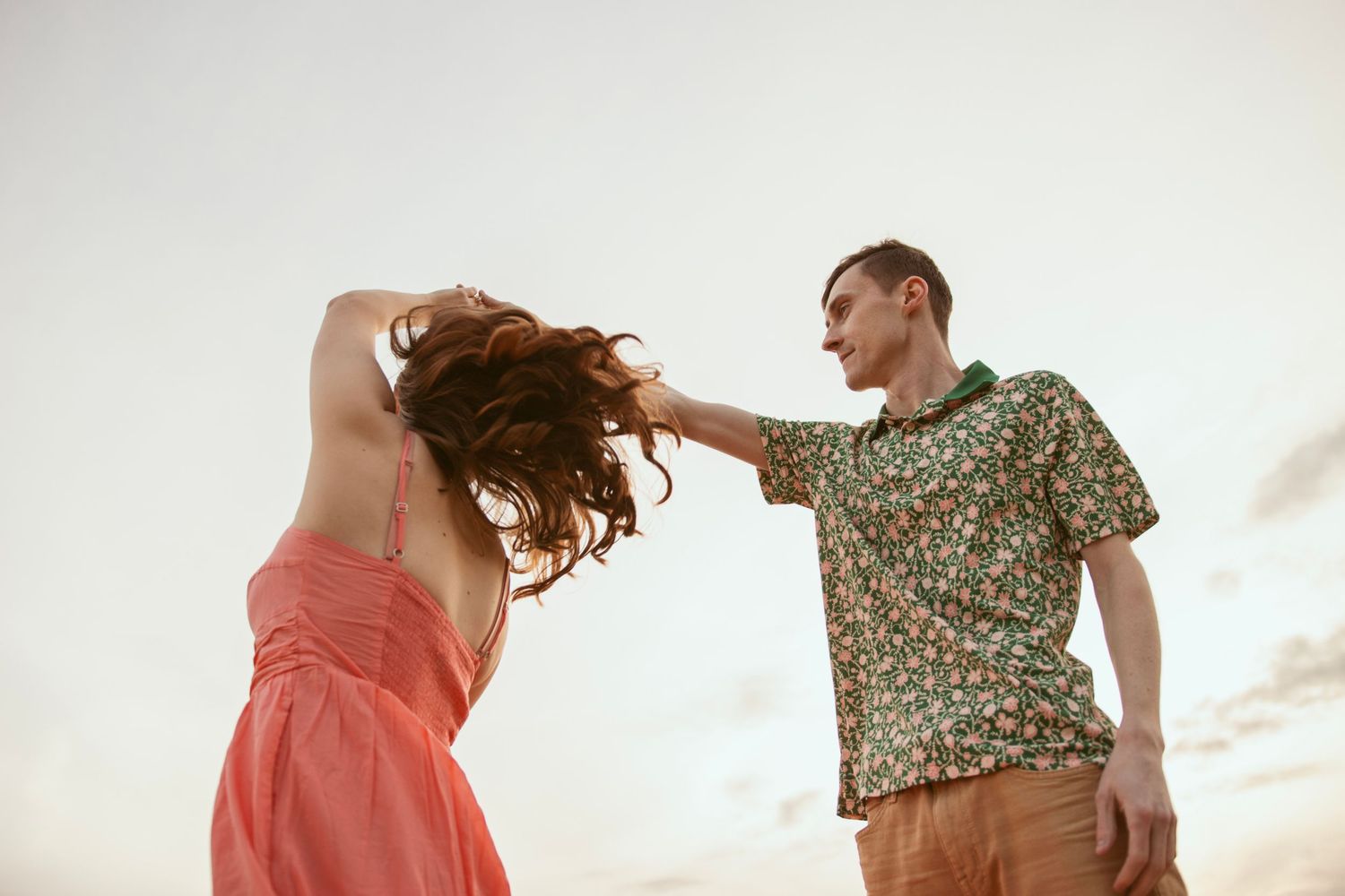 Playful couple dancing outdoors at sunset with her coral dress swaying in motion.