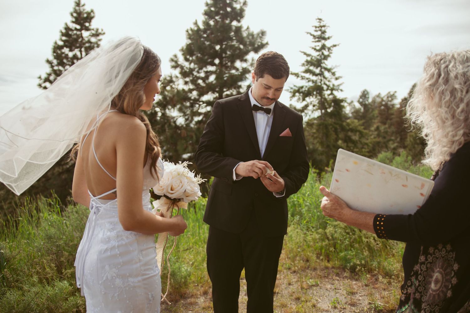 A wedding ceremony takes place outdoors with evergreen trees in the background as a couple exchanges vows.