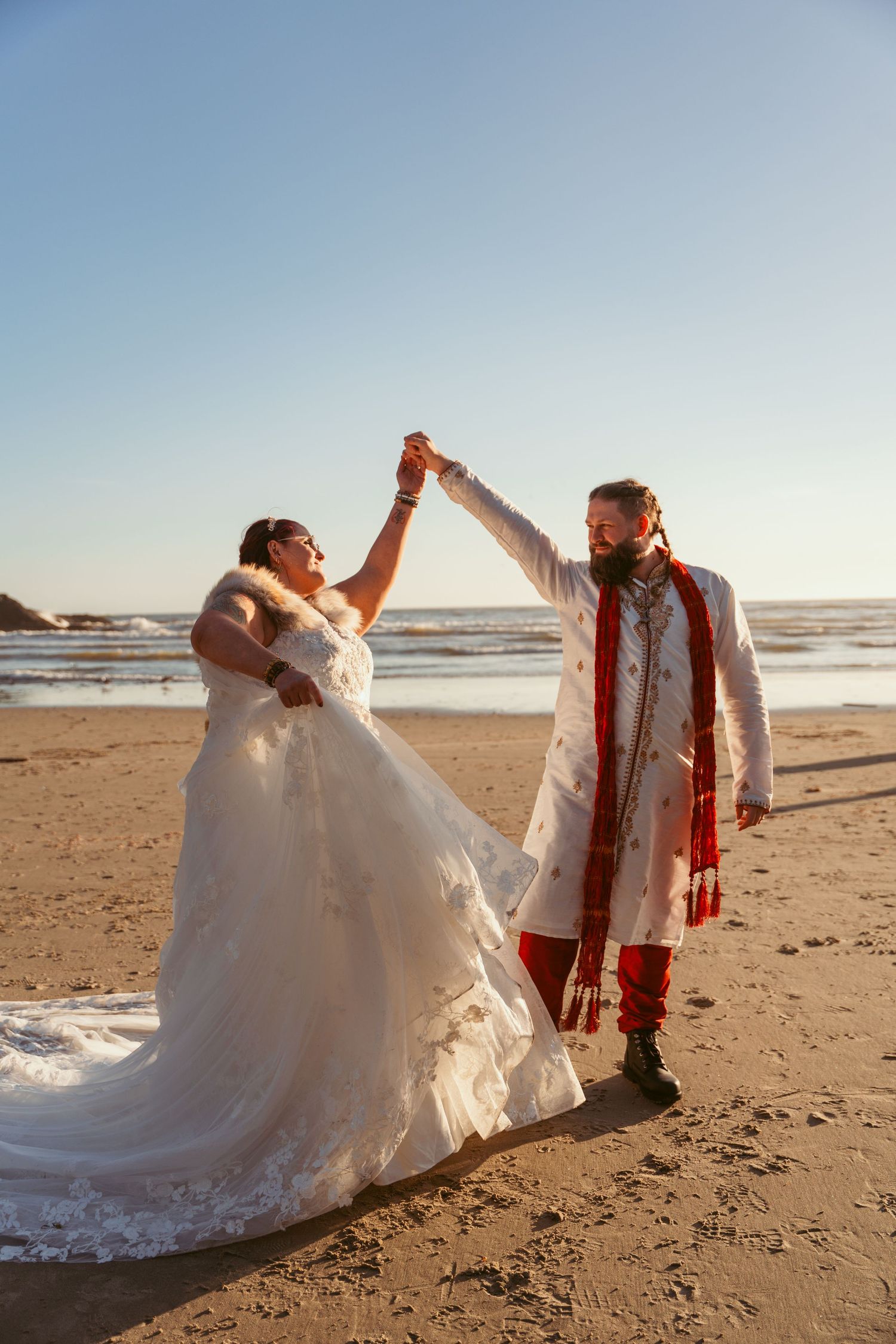 A couple poses dramatically on a beach at sunset in wedding attire.