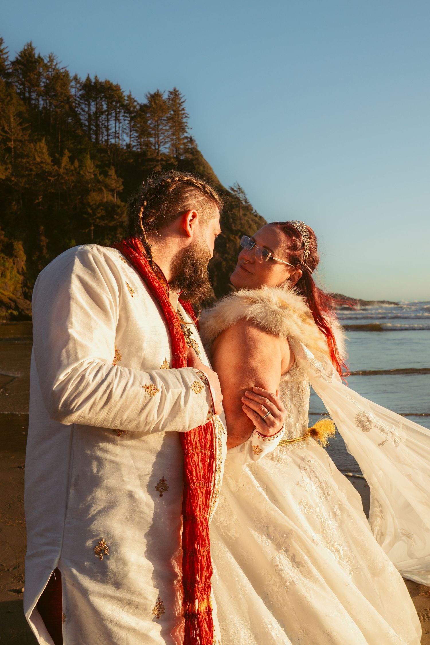 A couple in white wedding attire share a tender moment on a scenic beach at sunset.