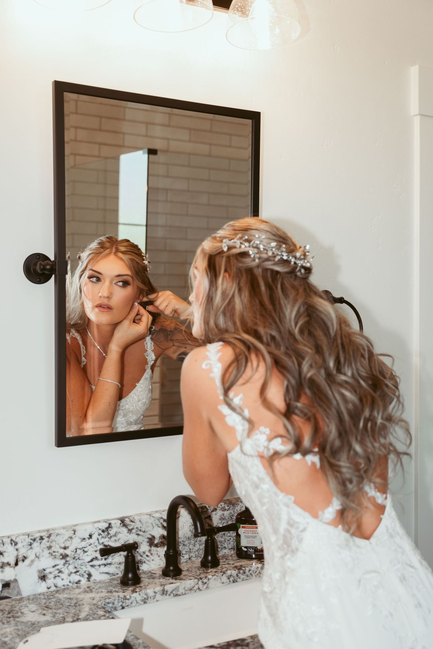 Bride adjusting jewelry while looking in bathroom mirror during wedding preparations.
