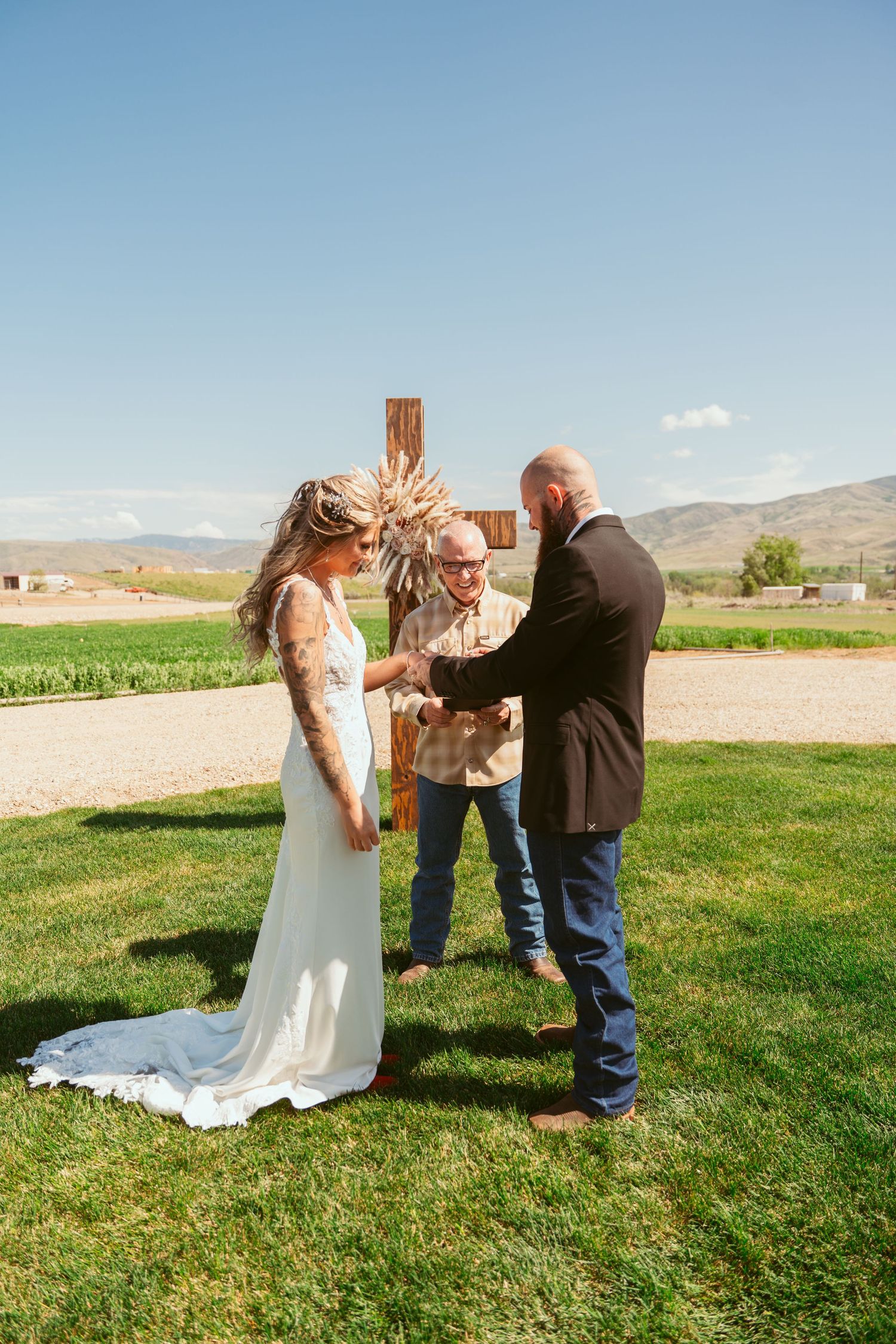 An intimate outdoor wedding ceremony takes place against a mountain backdrop with a rustic wooden cross.