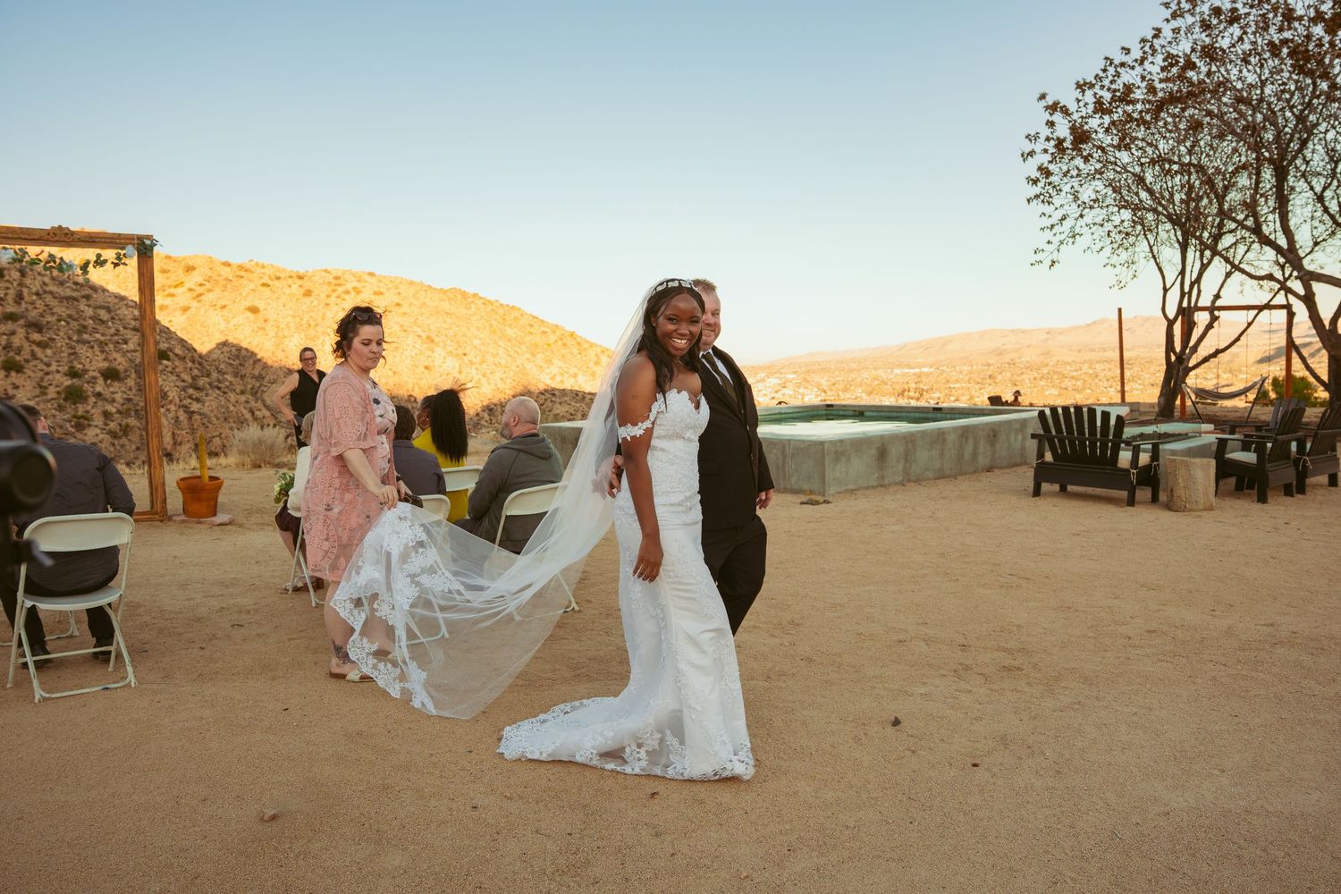 Desert wedding celebration with guests seated outdoors as sun sets over mountain landscape.