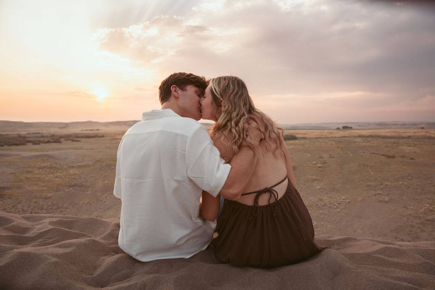 A couple sits close together on desert sand dunes during sunset.
