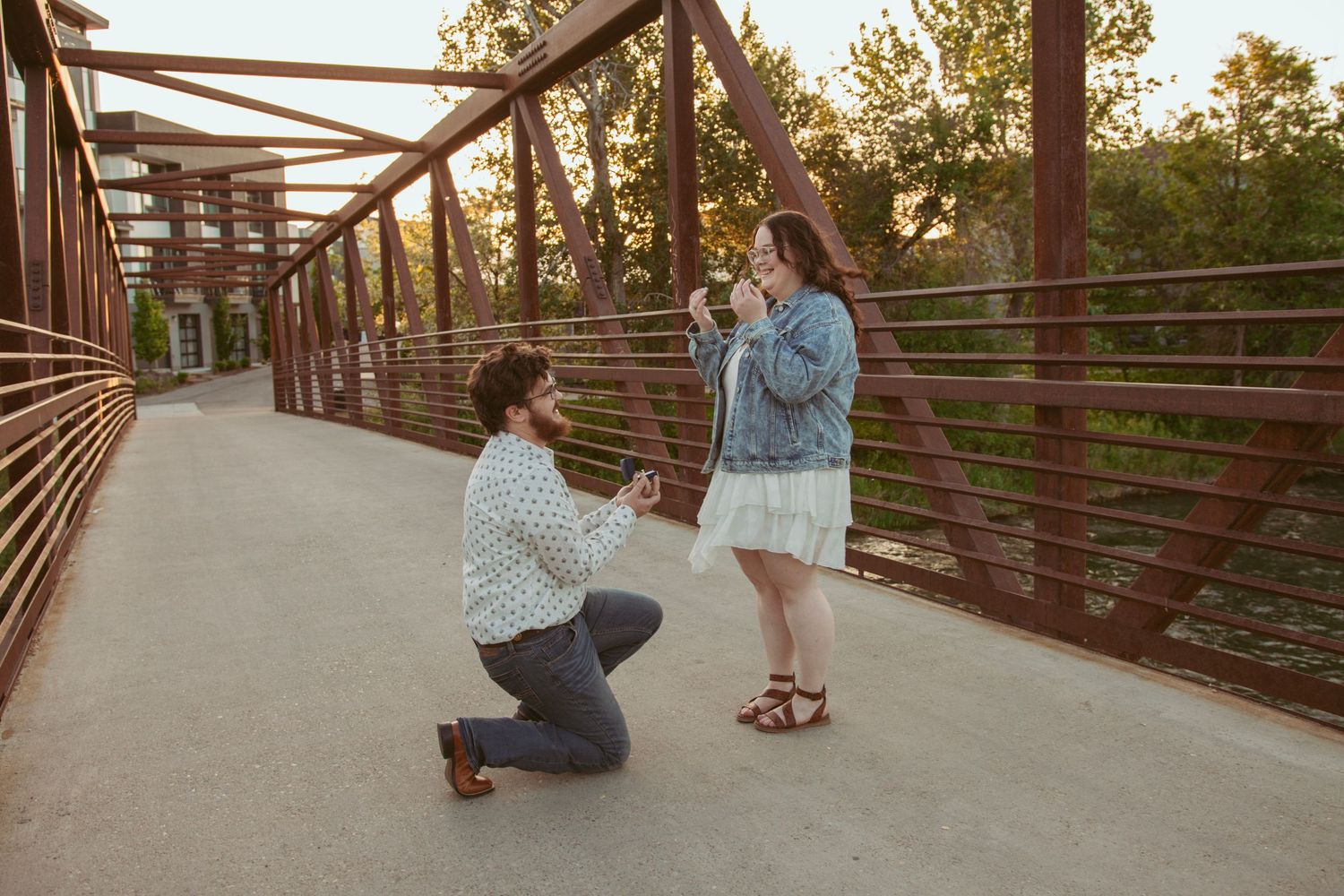 A romantic proposal scene unfolds on a rustic wooden bridge at sunset.