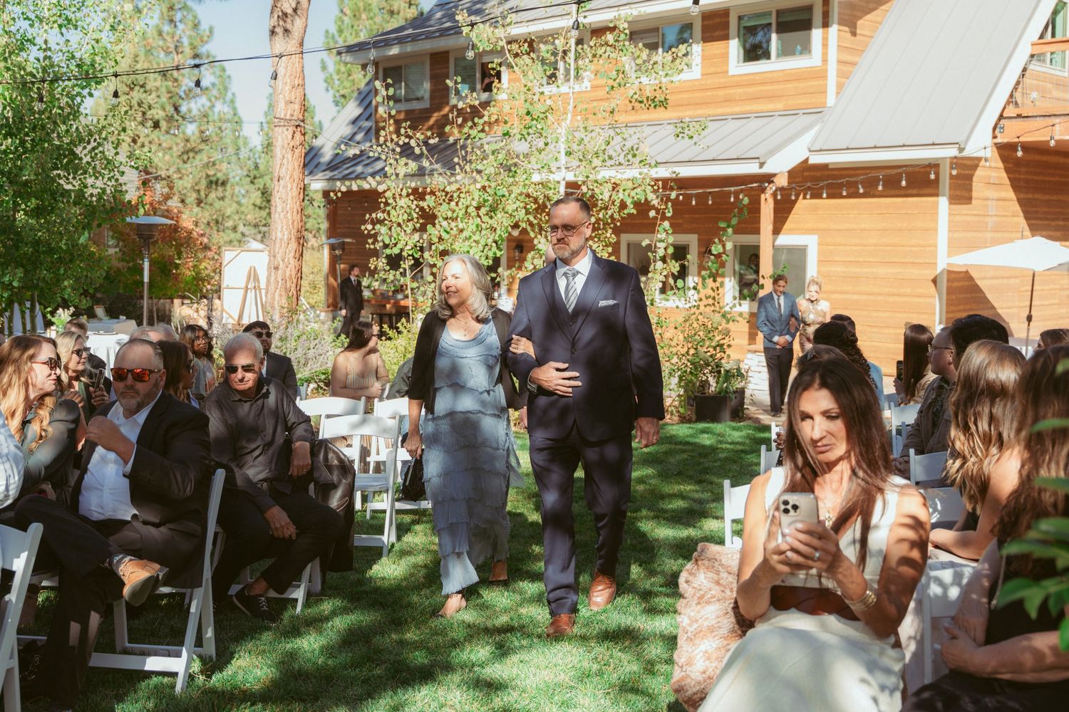 Guests seated on white chairs watch a wedding processional on a sunny outdoor lawn.