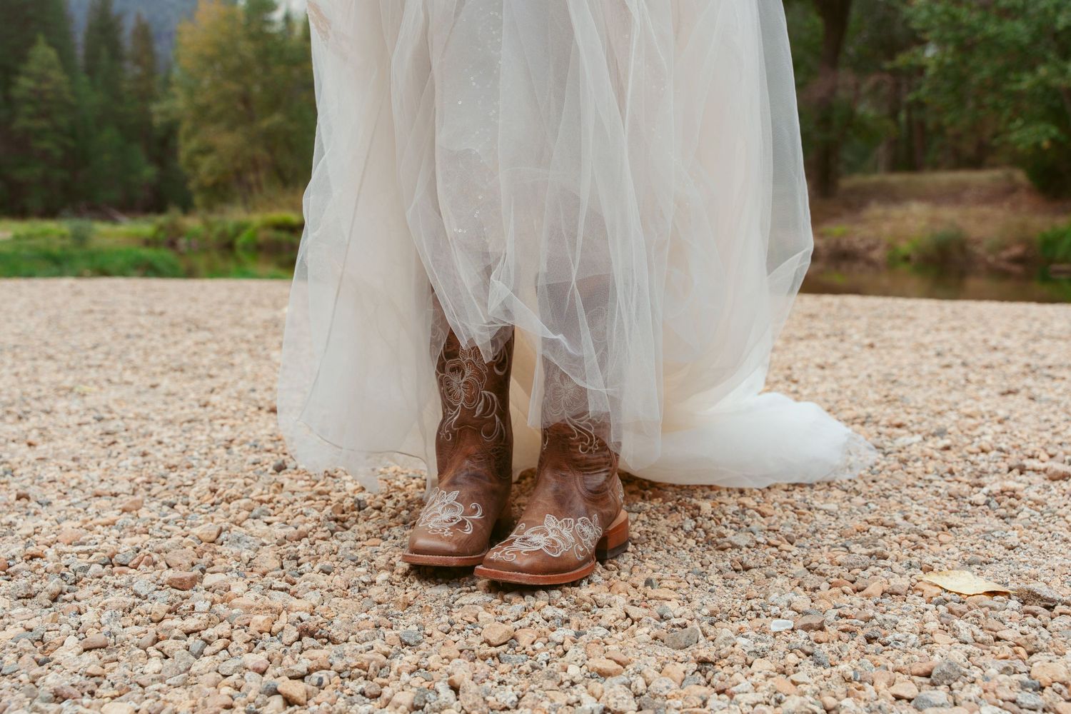 Brown cowboy boots peek out beneath a white wedding dress on a rocky riverbank.