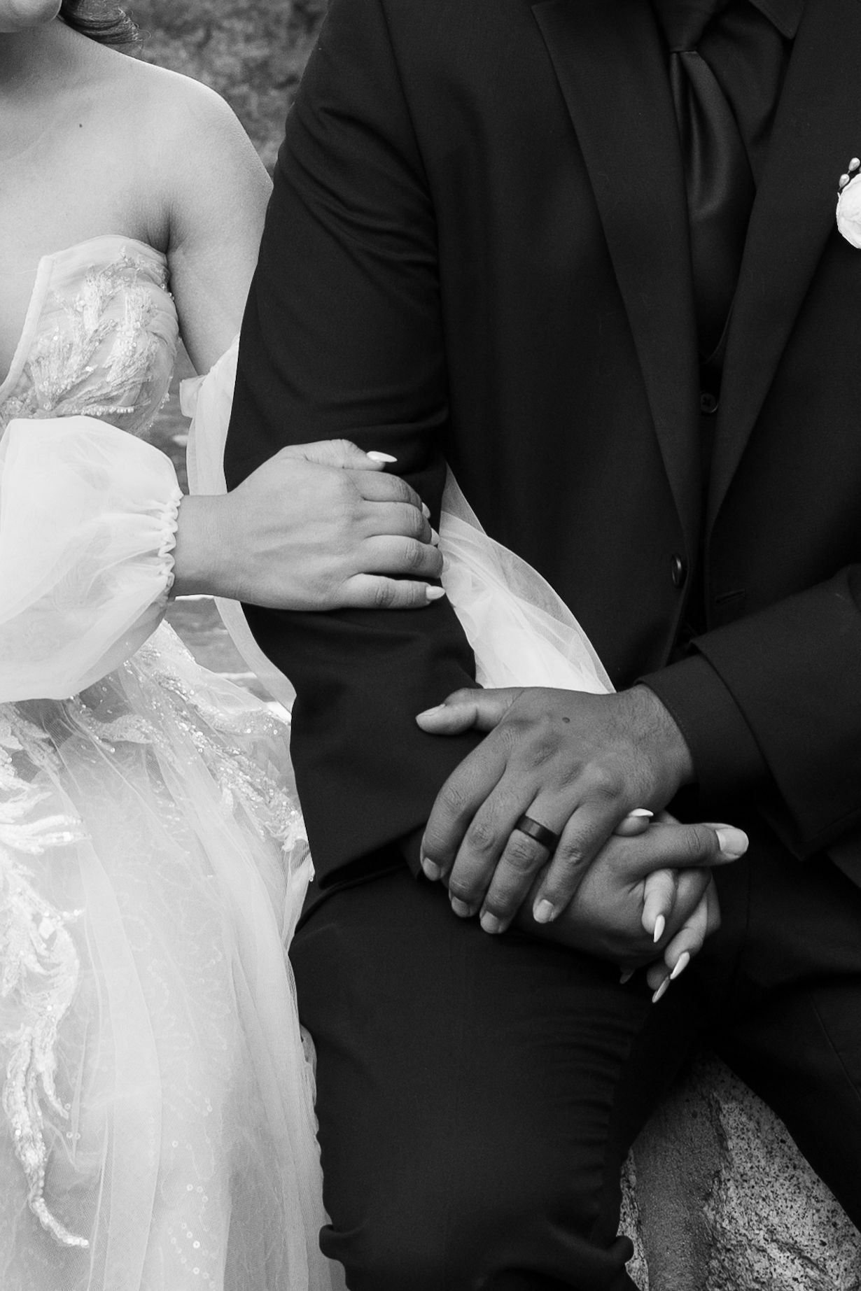 Close-up details of a bride in white lace and groom in black suit holding hands.