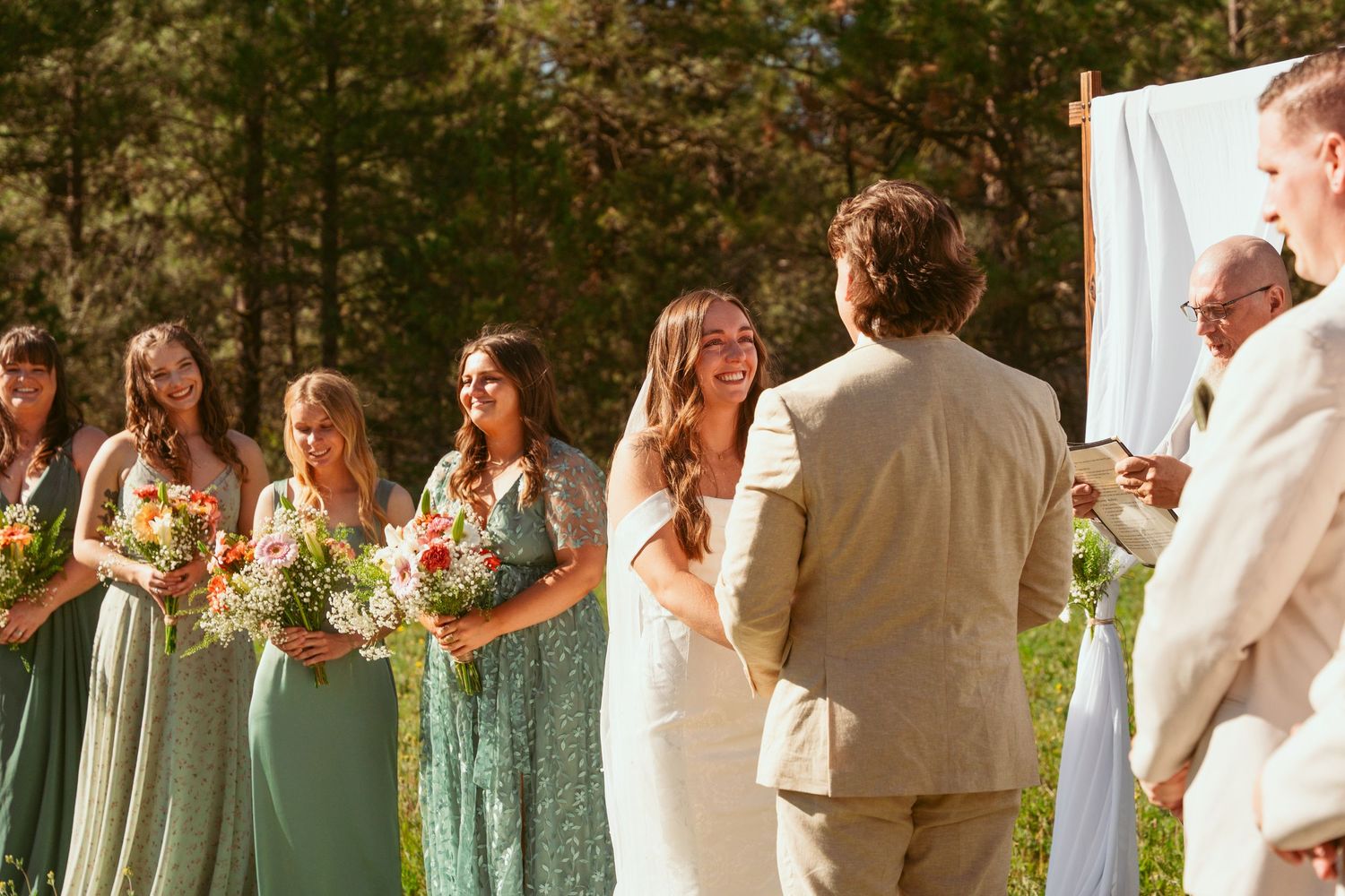 Wedding party members in light colored dresses and suits pose together in an outdoor setting.