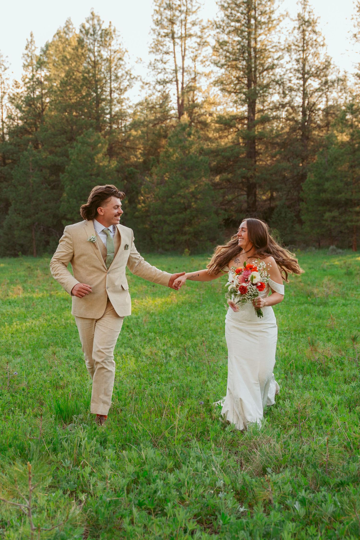 Wedding couple dances and spins playfully in a green meadow surrounded by pine trees.