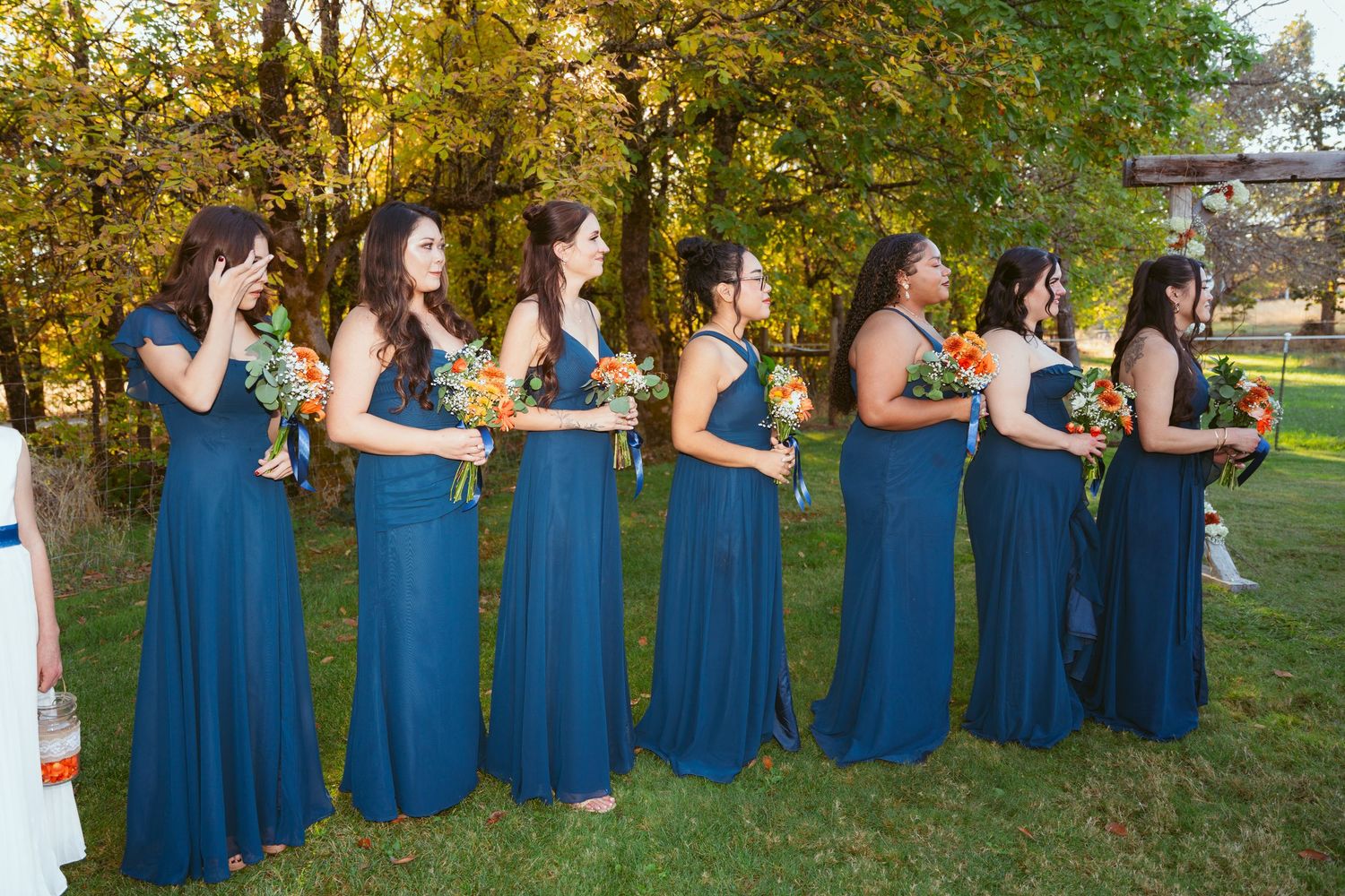 Row of bridesmaids in matching blue dresses stand together outdoors.