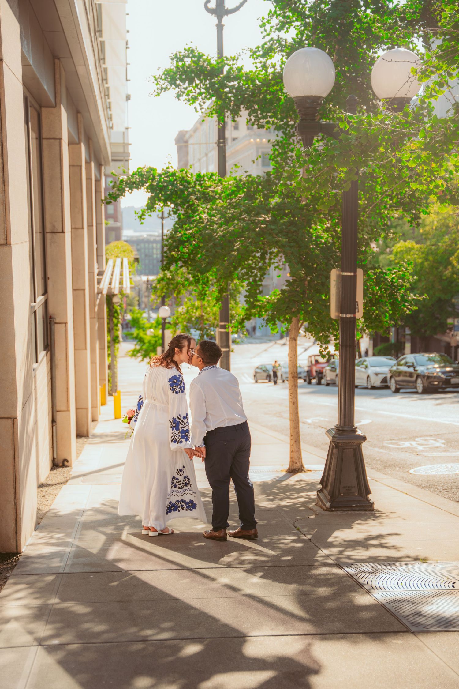 People gather on a sunny sidewalk with classic architecture and lamp posts.
