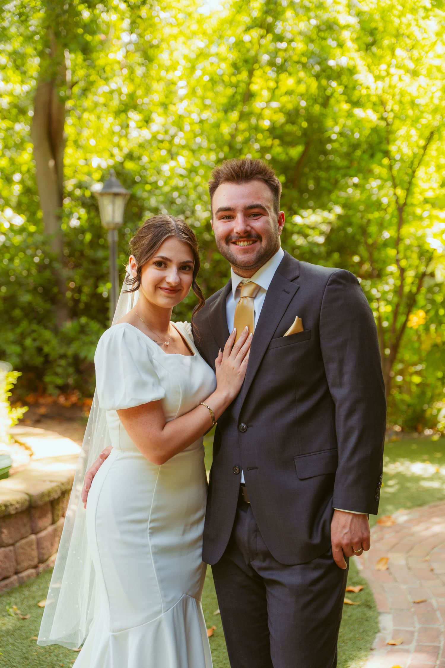 A wedding couple poses outdoors against a backdrop of green trees and brick retaining wall.