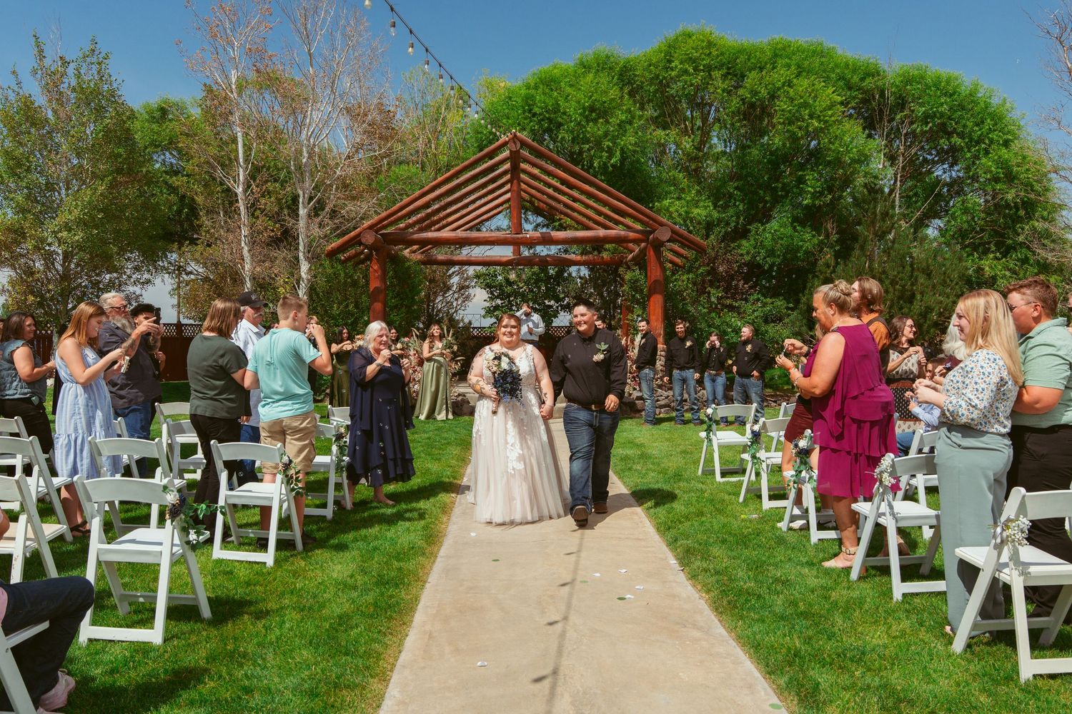 Outdoor wedding ceremony setup with white chairs and wooden pergola on a grassy lawn under blue skies.