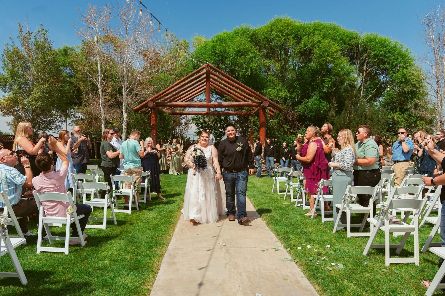 Guests seated in white chairs for an outdoor wedding ceremony with wooden pergola and green landscaping.
