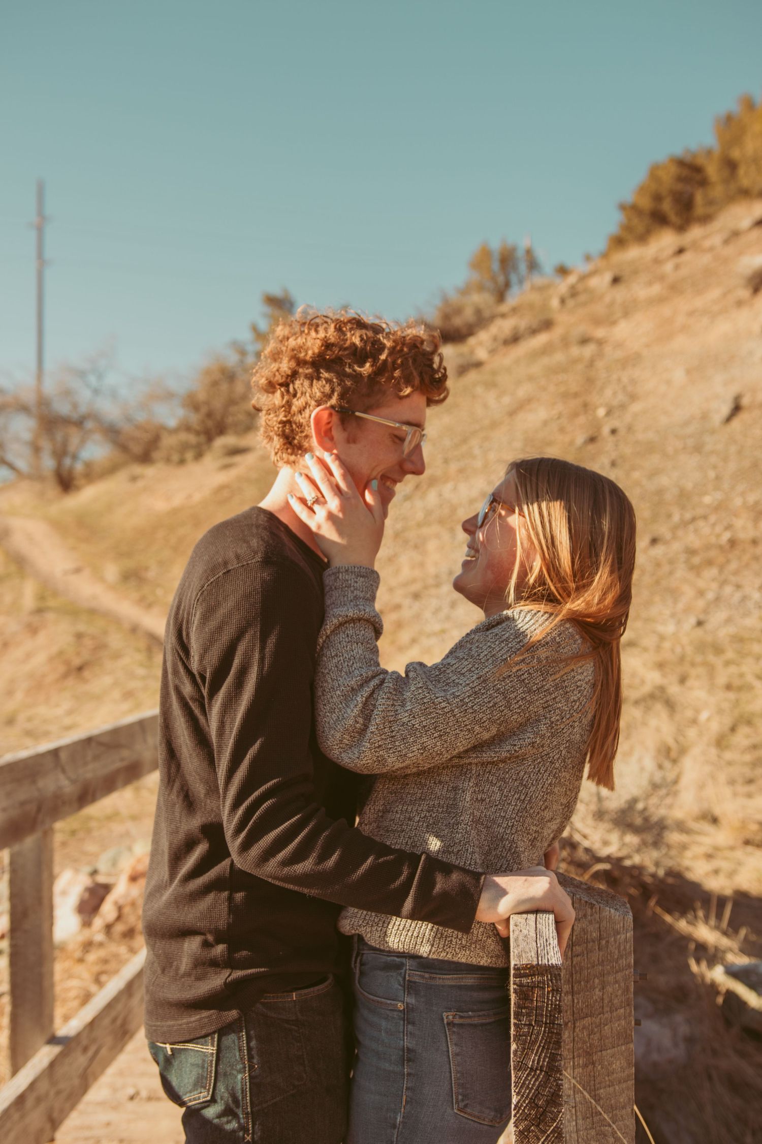 A couple in casual attire embraces against a rustic wooden fence during golden hour on a hillside.