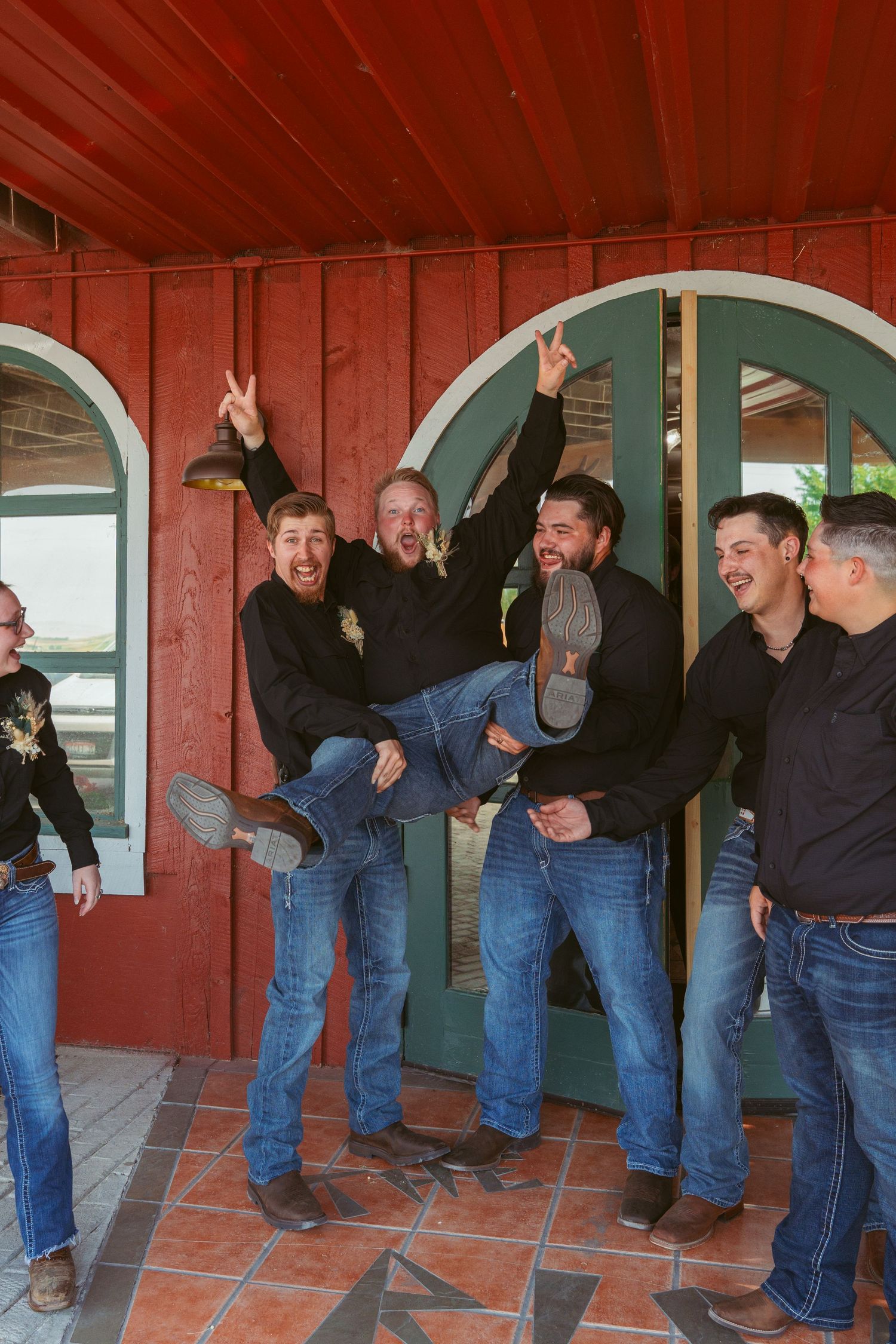 Groomsmen in jeans and dark shirts strike funny poses in front of red barn with arched doorways.
