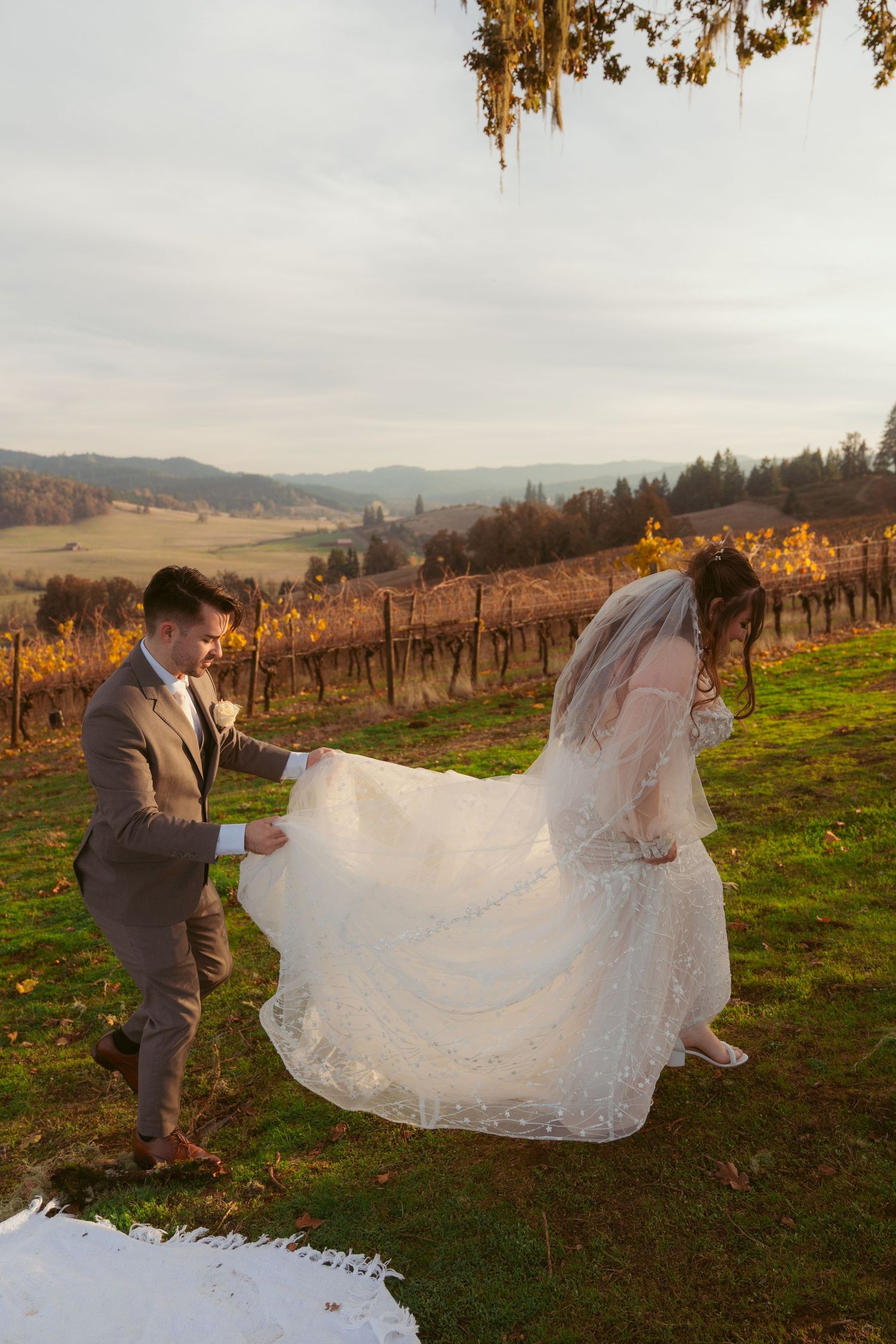 Wedding couple playfully twirls the bride's dress in a scenic vineyard setting at sunset.