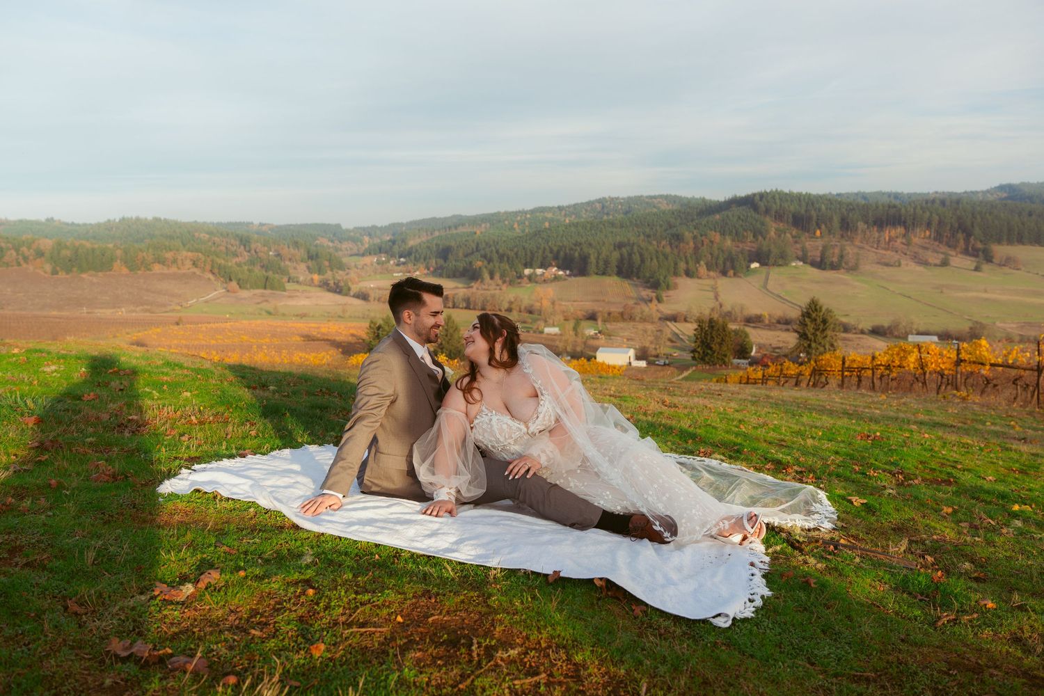 Couple in wedding attire laying on white fabric against scenic mountain backdrop.