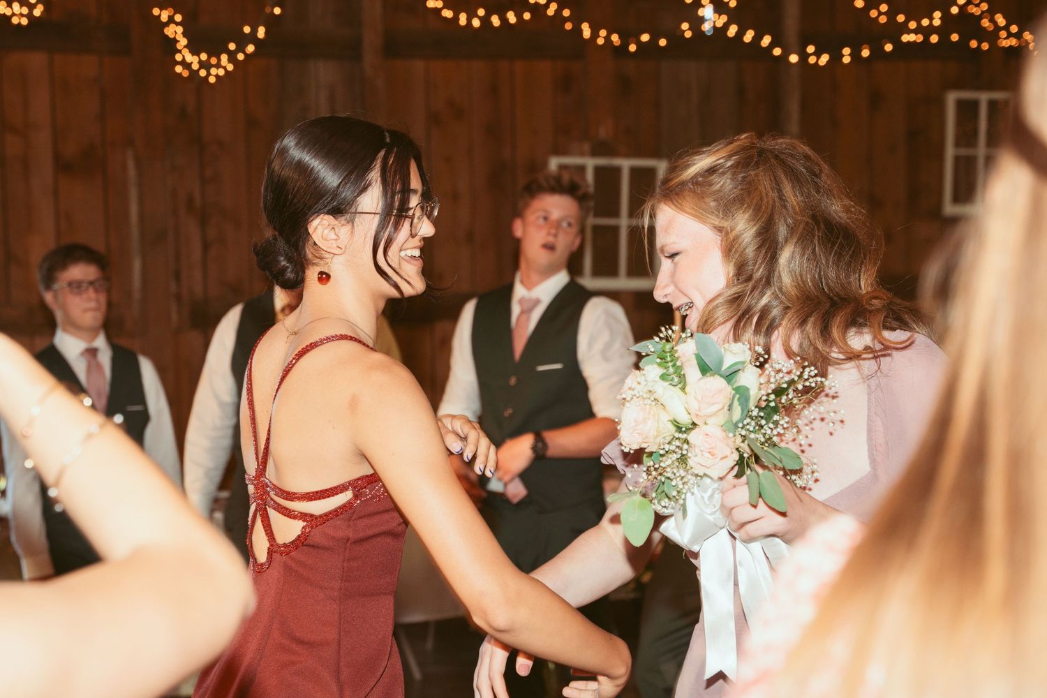 Festive wedding reception with dancing guests under glowing string lights in rustic venue.
