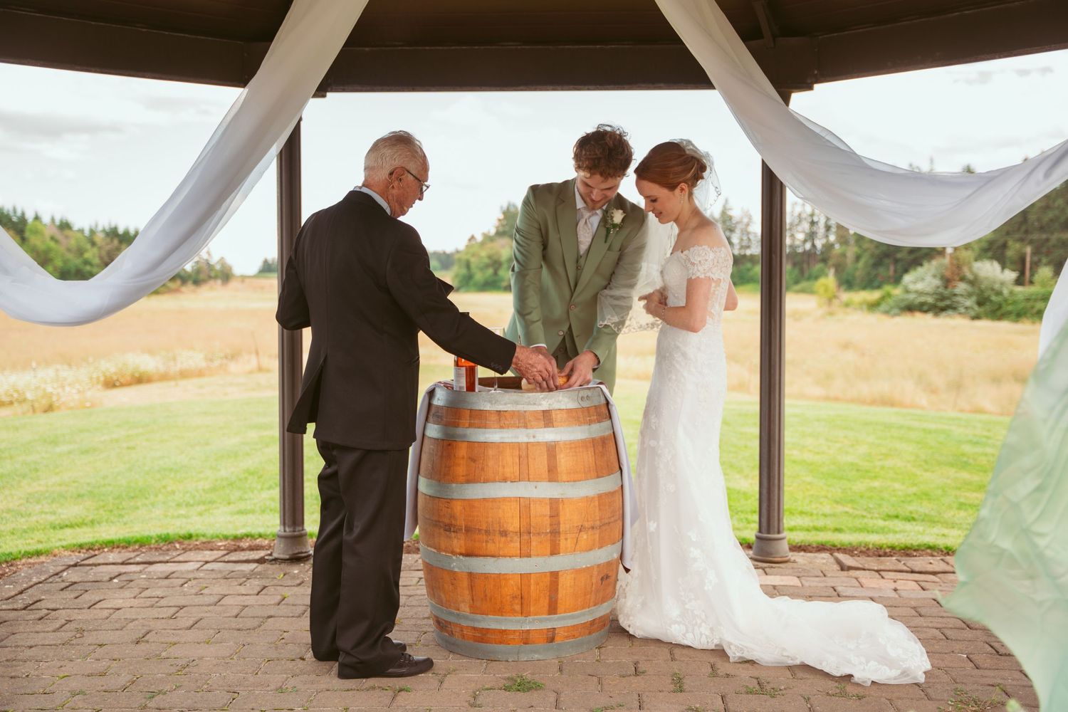 Wedding ceremony under a wooden pavilion with wine barrels and white fabric draping.