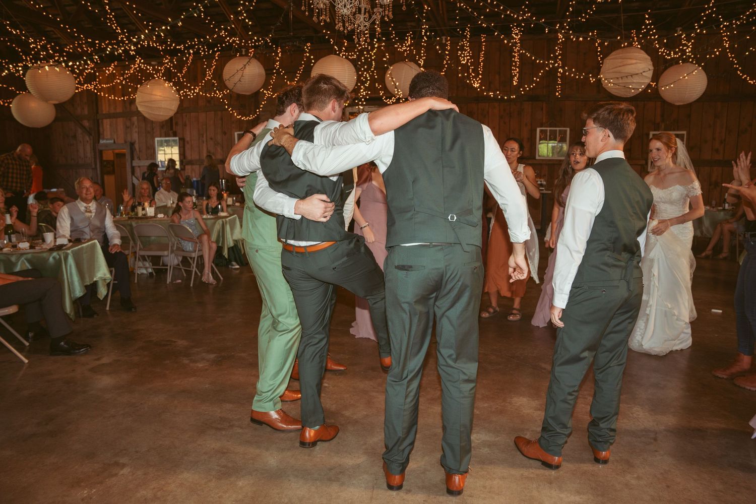 Wedding guests dance in coordinated outfits under warm string lights in a rustic barn setting.