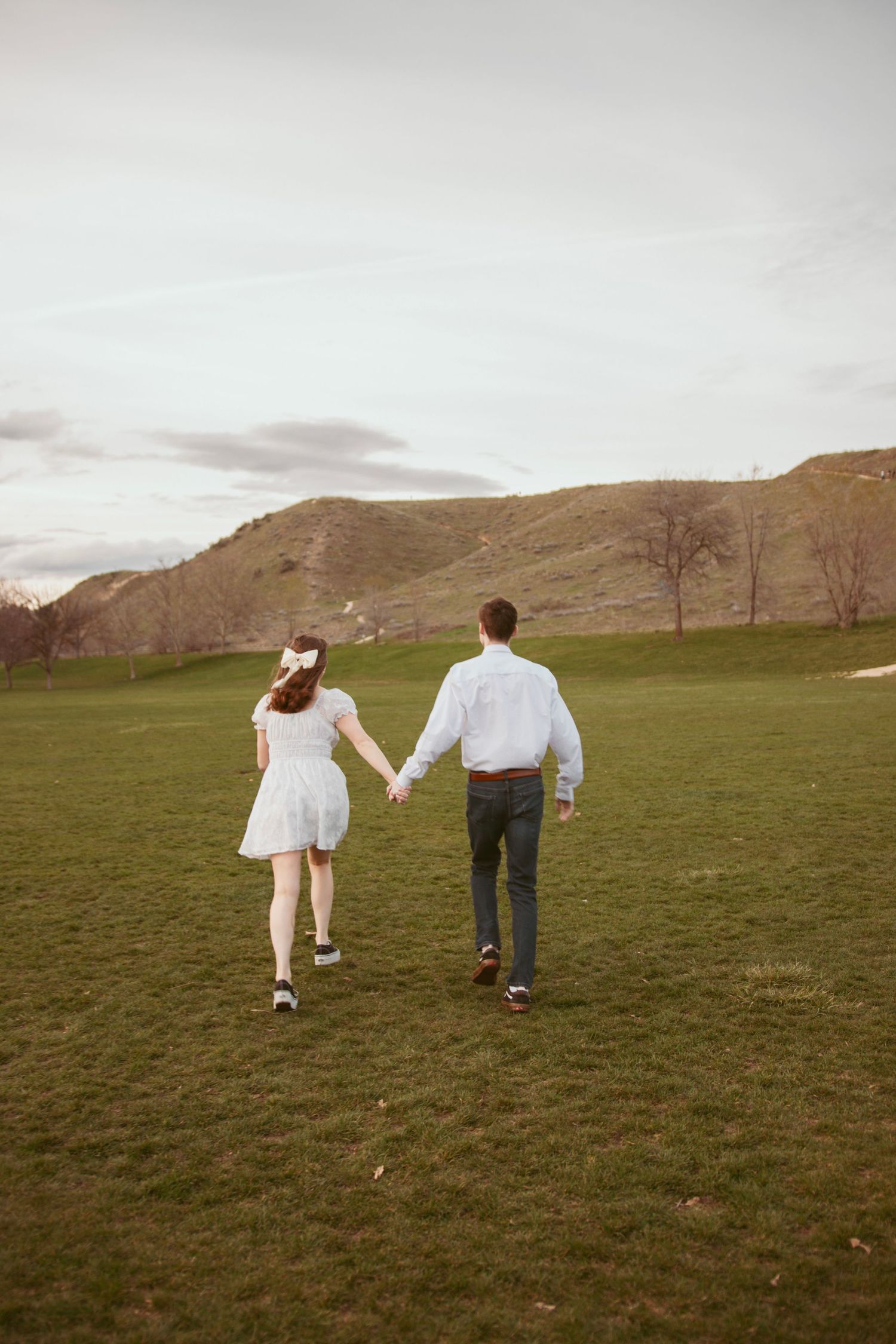 A couple walks hand in hand across a scenic green field with rolling hills in the background.