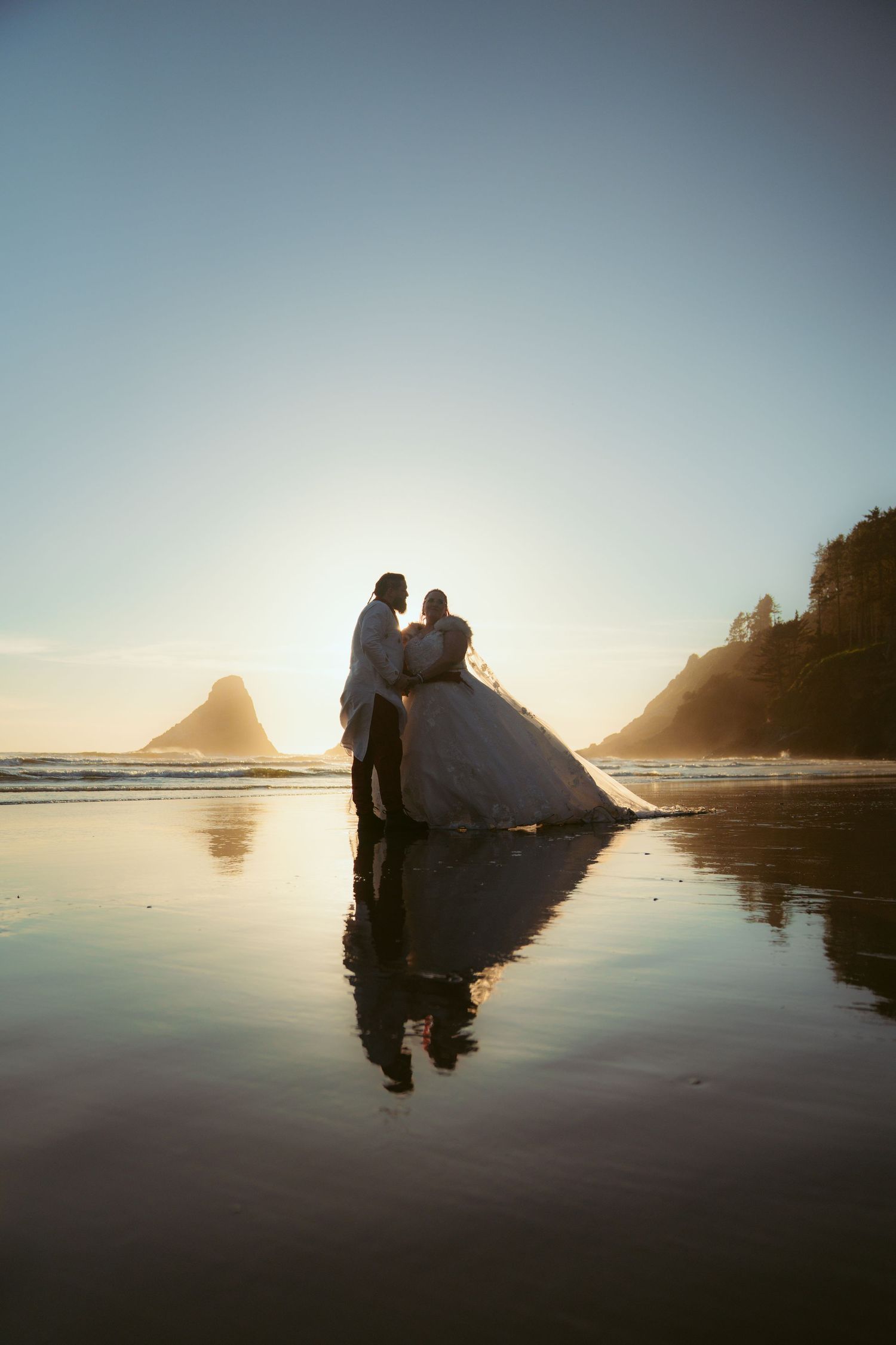 Silhouetted figures standing on mirror-like beach with mountains and sunset in the distance.
