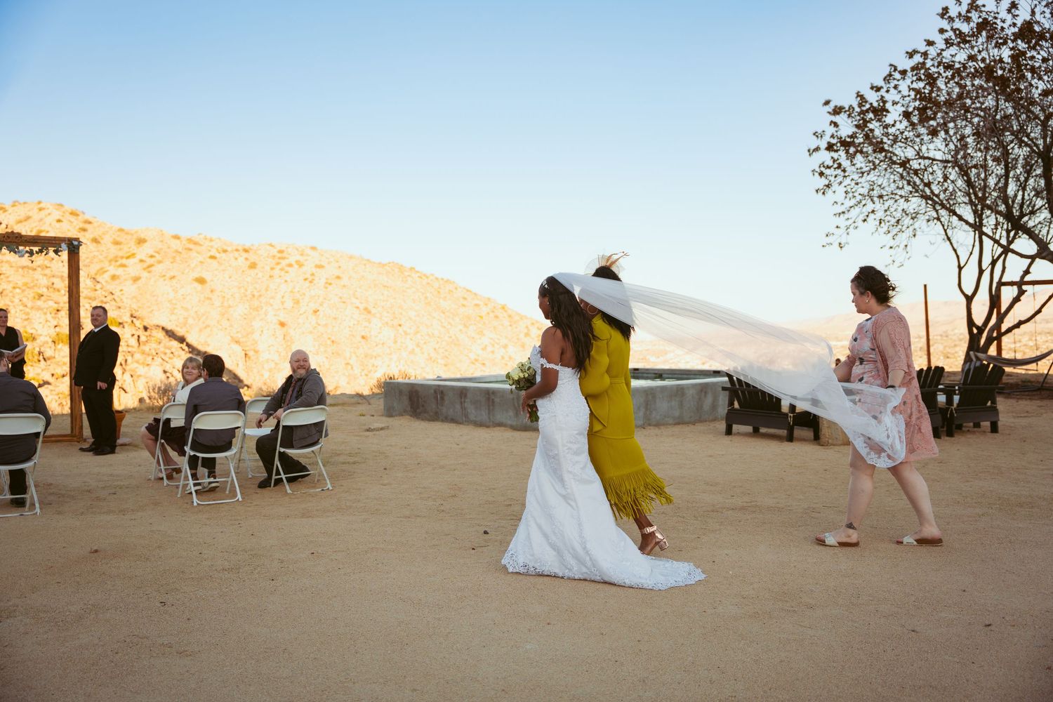 Wedding guests and ceremony setup in desert landscape with golden sunset lighting.