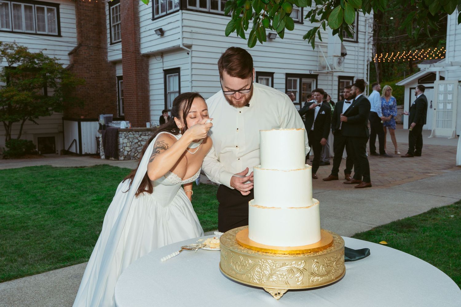 Newlyweds share a laugh while cutting their elegant white tiered wedding cake outdoors.