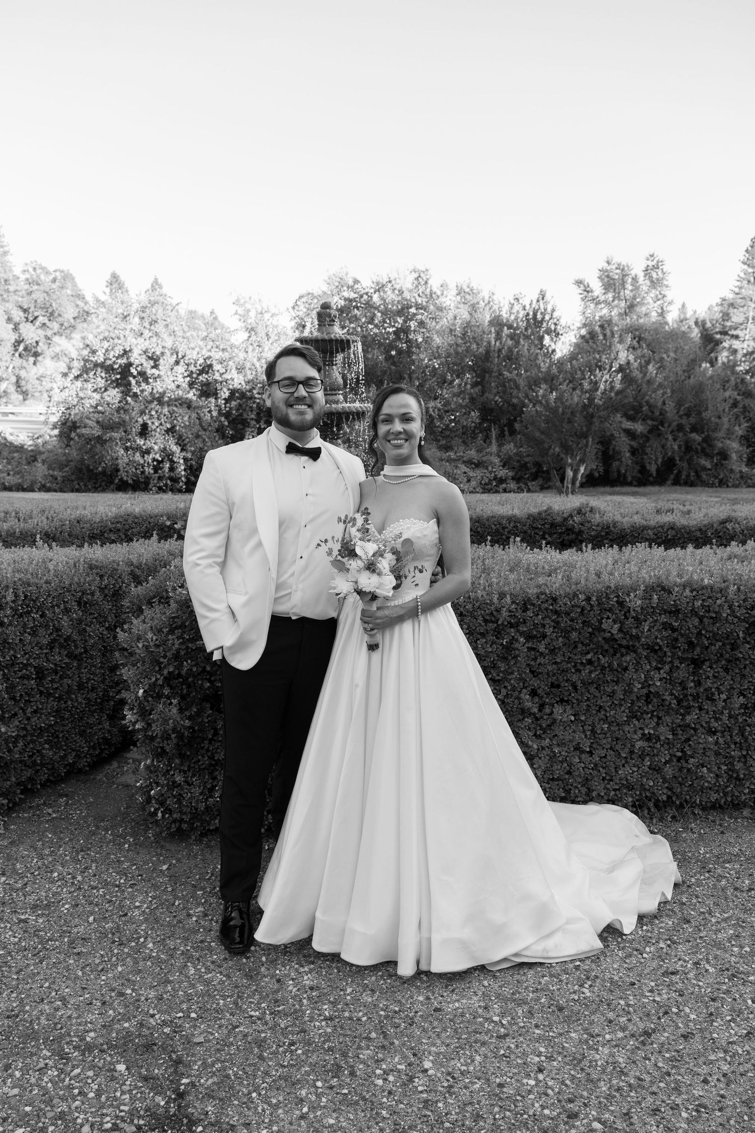 A wedding couple poses together in formal attire against a garden backdrop in black and white.