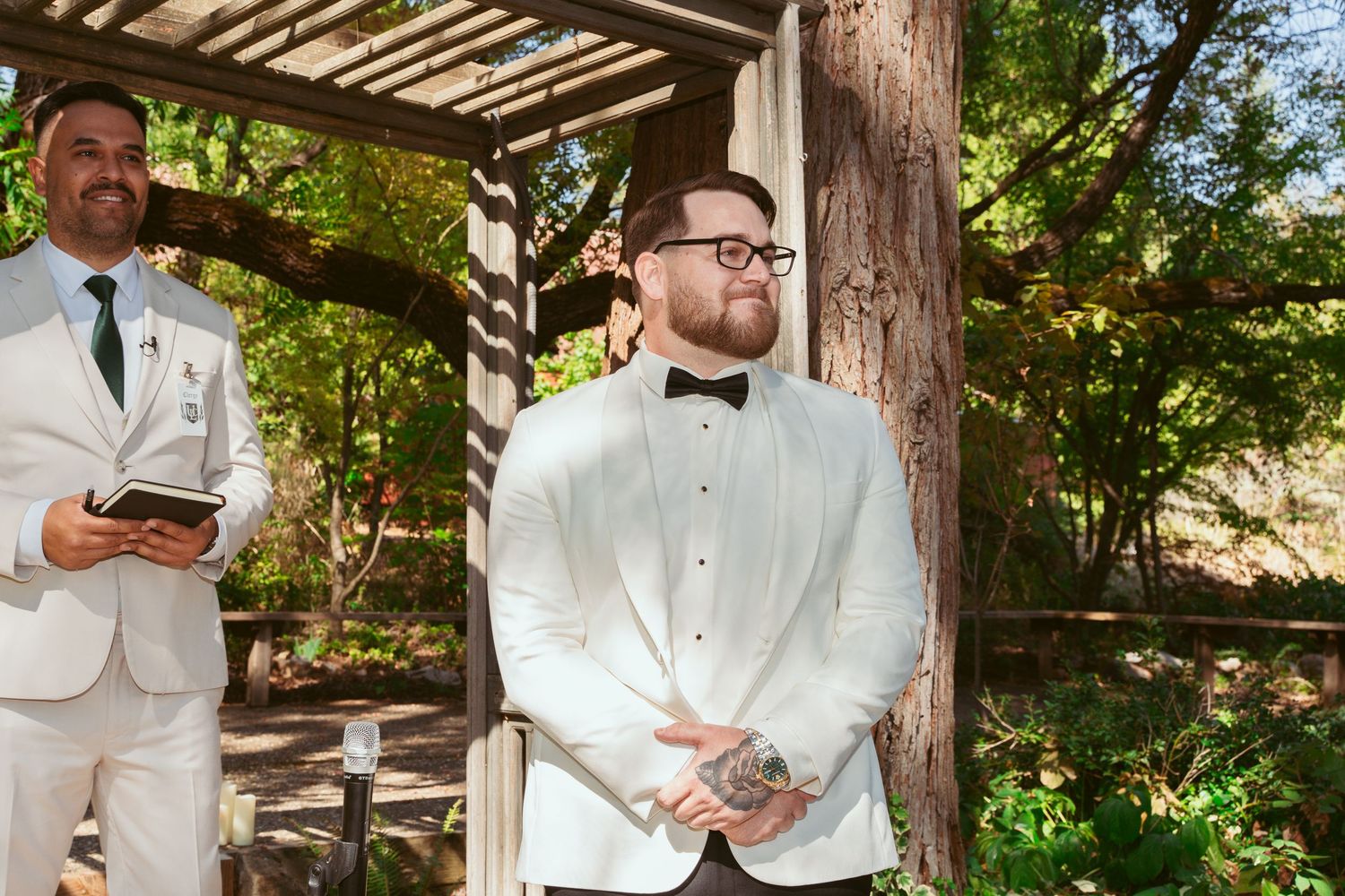 Groomsmen in formal attire pose under a wooden pergola during outdoor wedding.