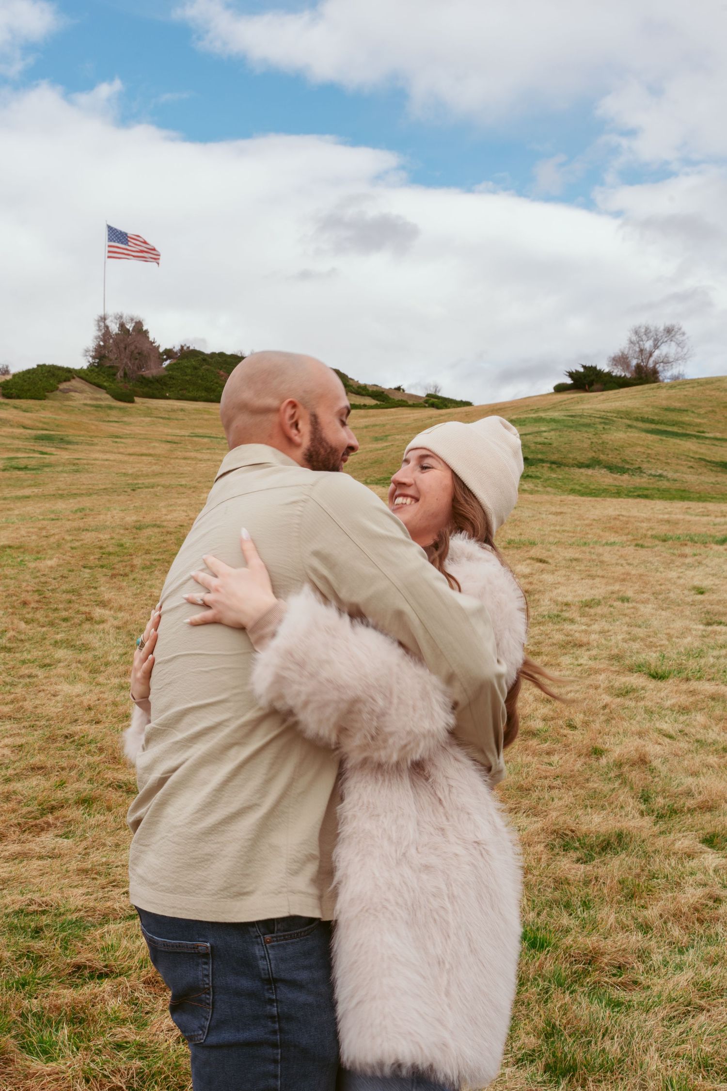 Couple in casual wear embracing on grassy hillside with American flag in distance.