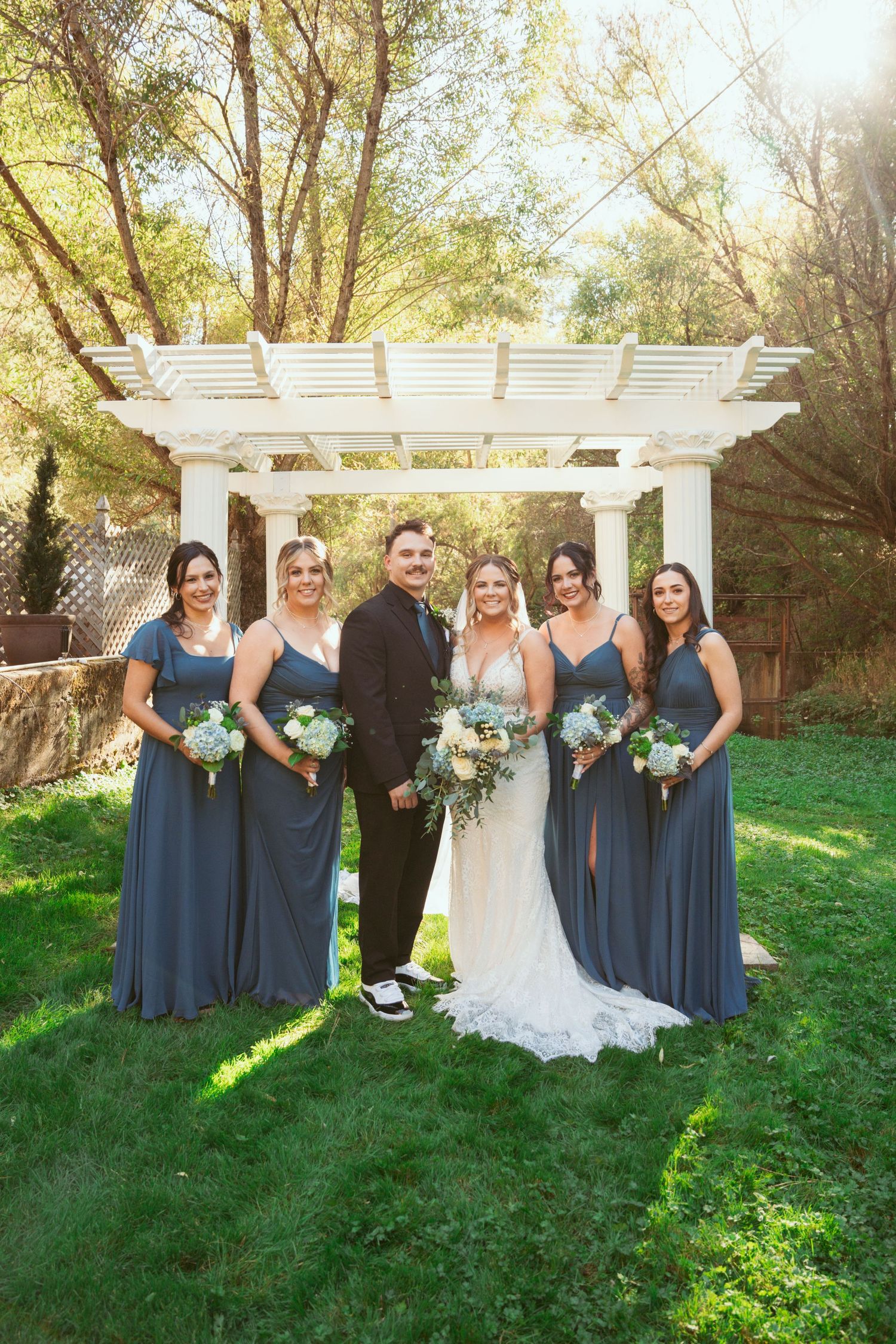 Wedding party members line up for photos under a white garden pergola on a sunny afternoon.